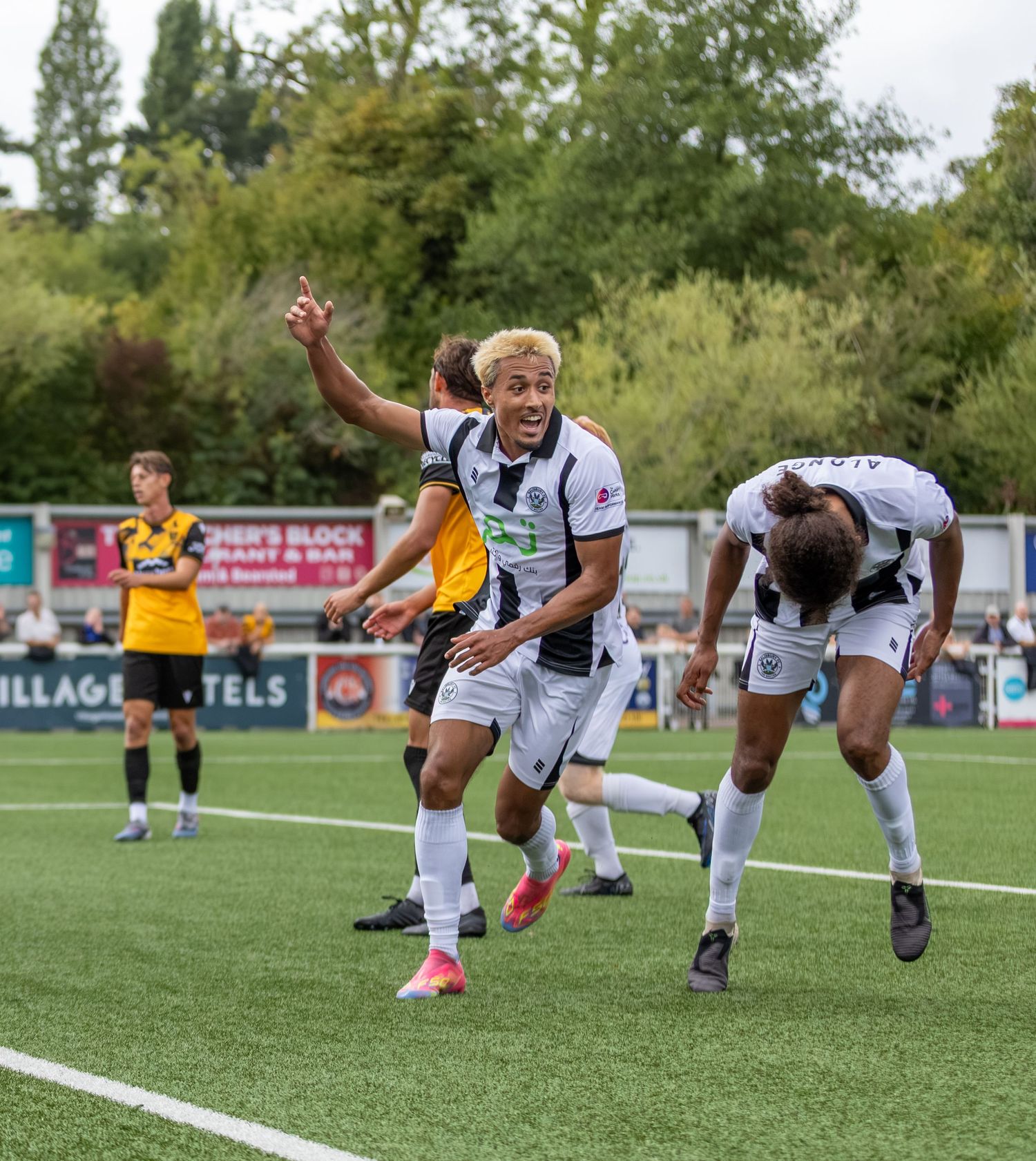 Soccer player celebrates goal in white kit, raising arm triumphantly on green pitch during competitive match.