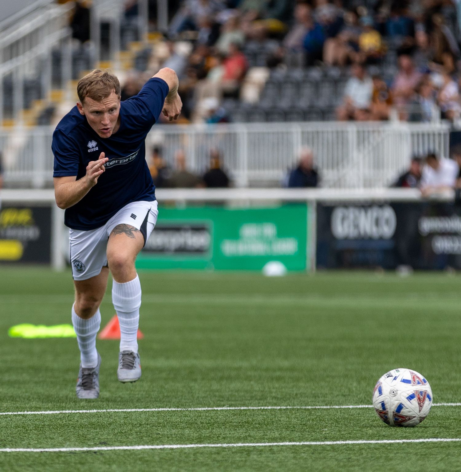 Soccer player in navy kit kicking ball on green turf during a competitive match at a stadium.