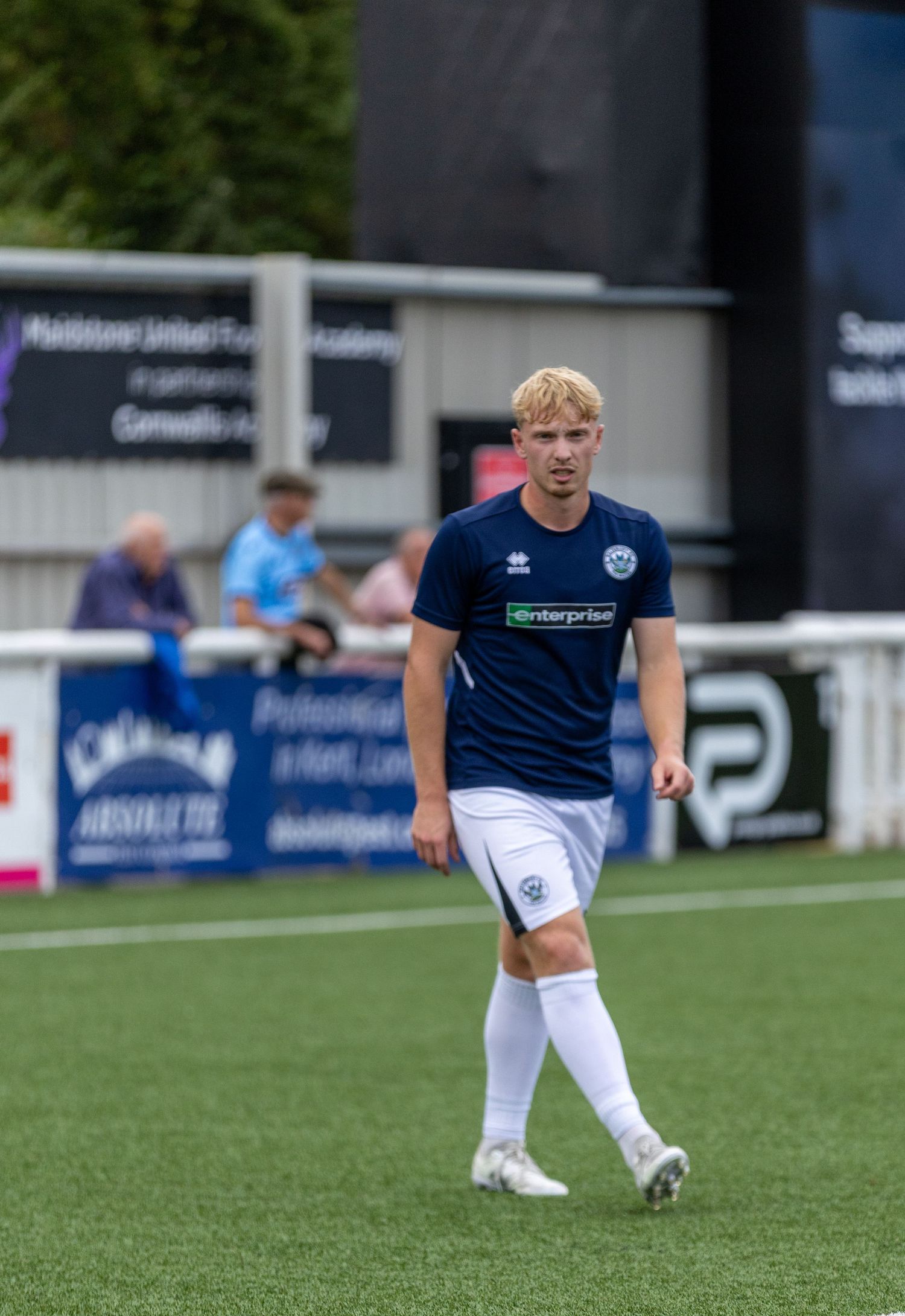 A young male soccer player in a navy blue kit walks across a green pitch during a match.