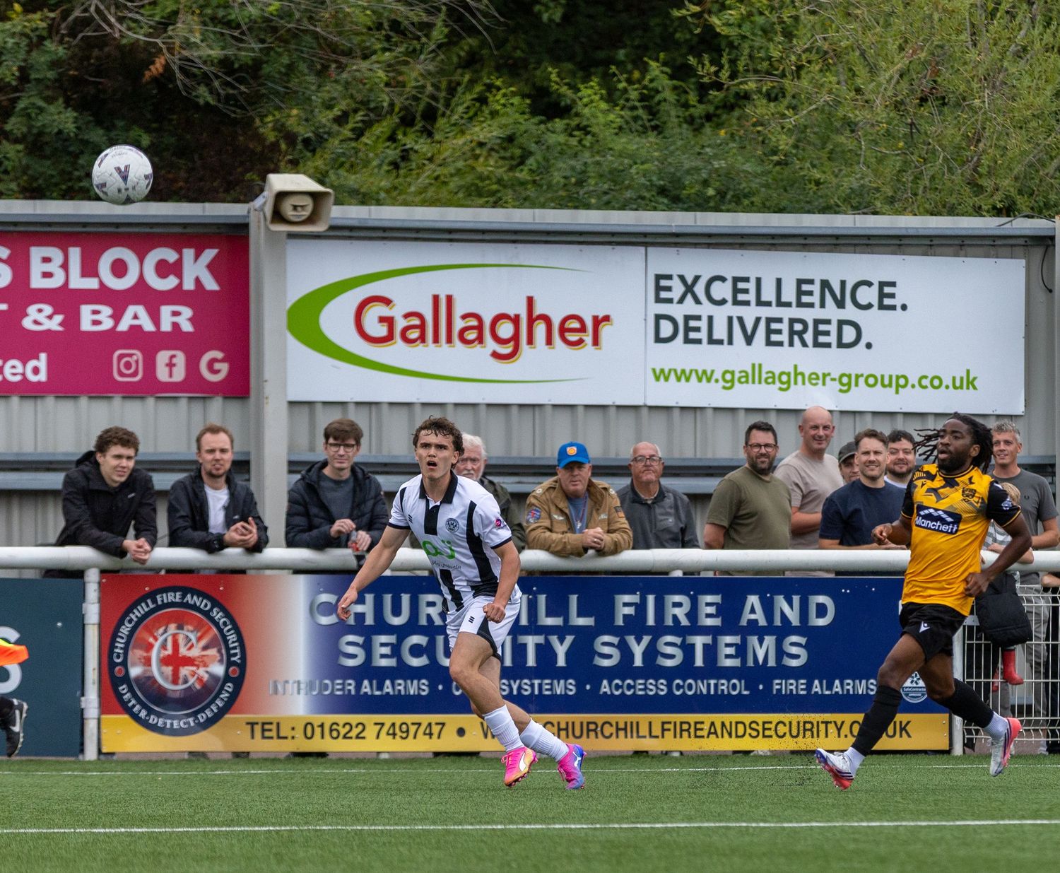 Two soccer players compete for the ball during a match near advertising boards for Gallagher and Churchill Fire and Security Systems.