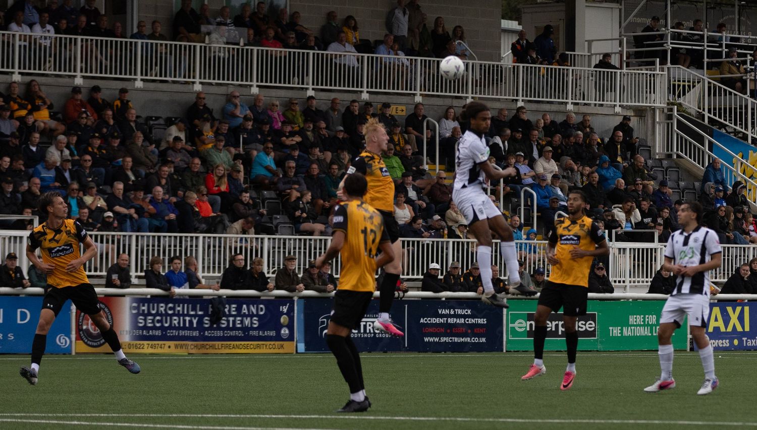 Soccer players compete for a header during a match, with one player leaping above opponents in a stadium setting.