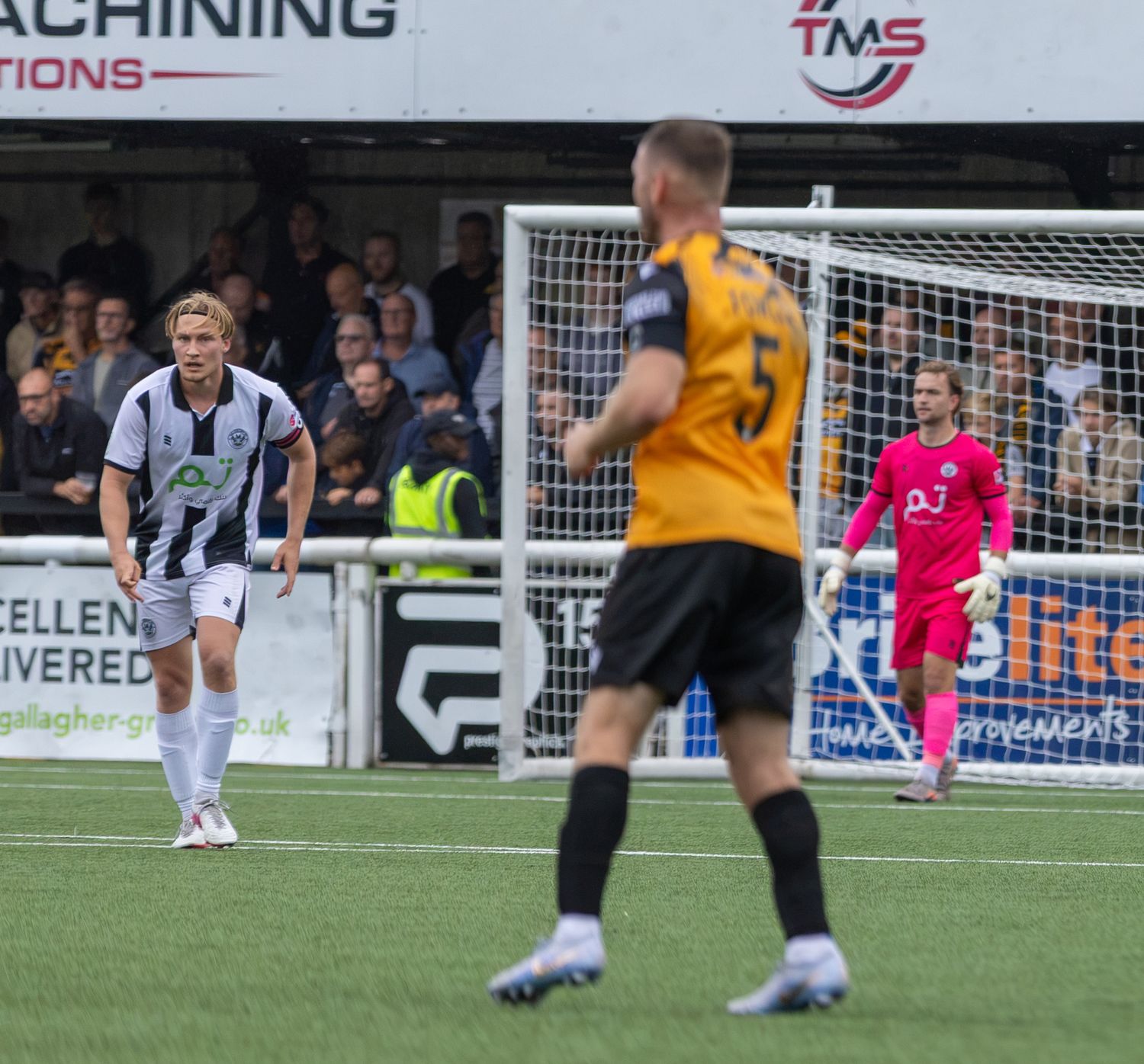 Football players on pitch during a match, with goalkeeper in pink kit and outfield players in black-white and amber strips.