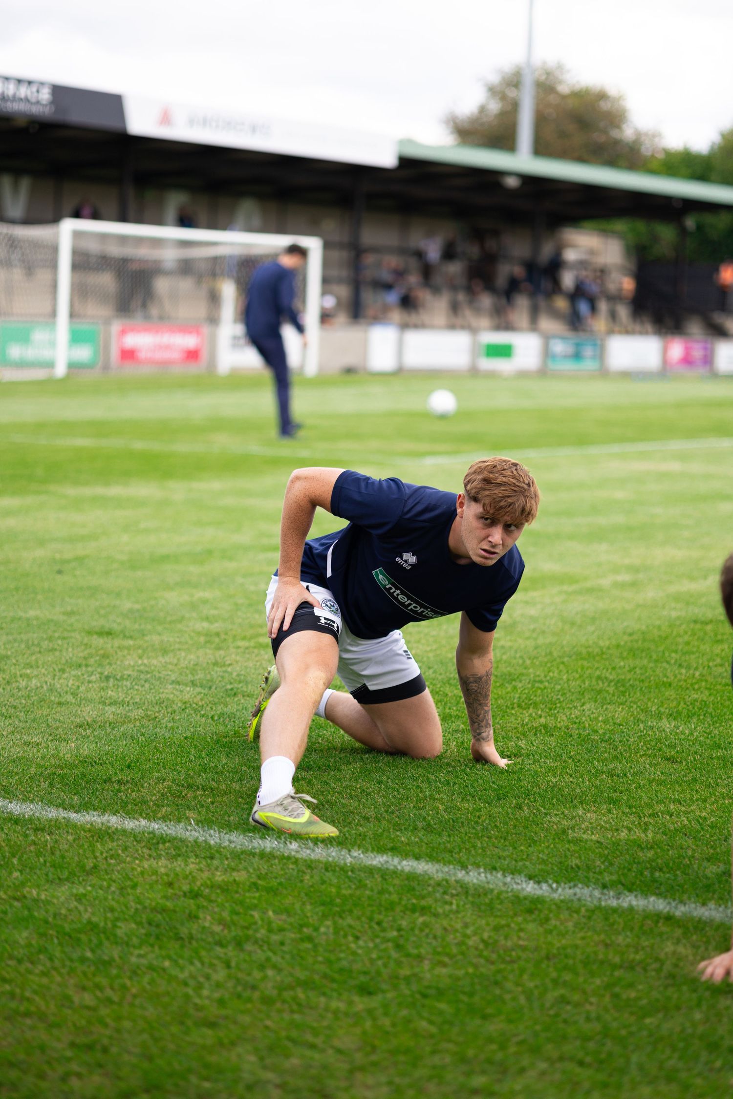 Young footballer stretching on grass pitch during pre-match warmup at a non-league football ground.
