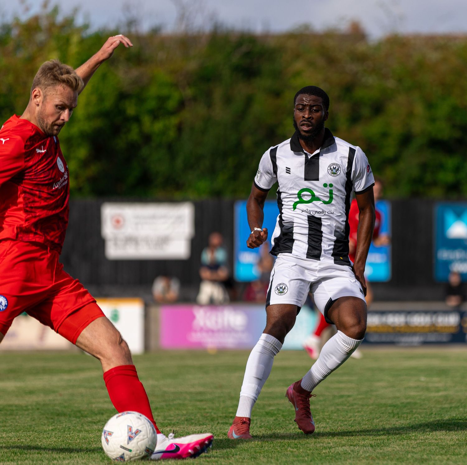 Two soccer players compete during a match, one in red and one in black and white striped kit, on a sunny green pitch.