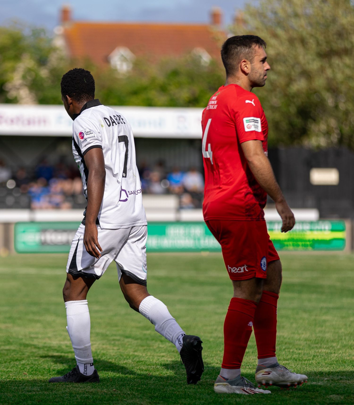Two soccer players in white and red jerseys walk on a grass field during a sunny match.