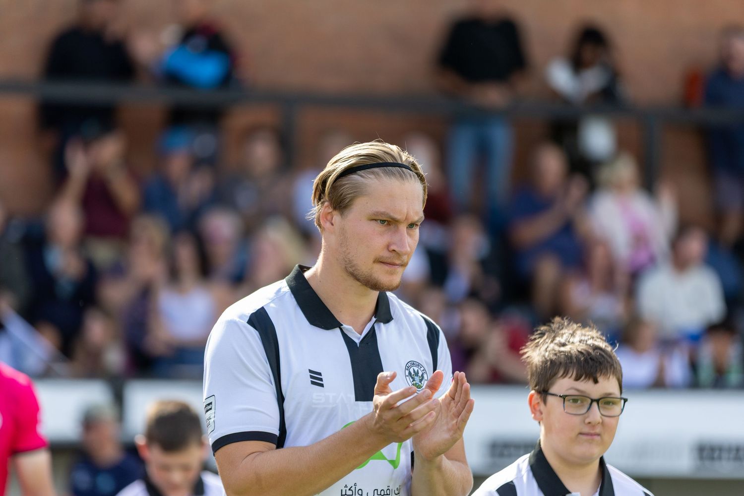 A footballer in black and white kit applauds fans at a match, with a young boy and crowd visible behind him.