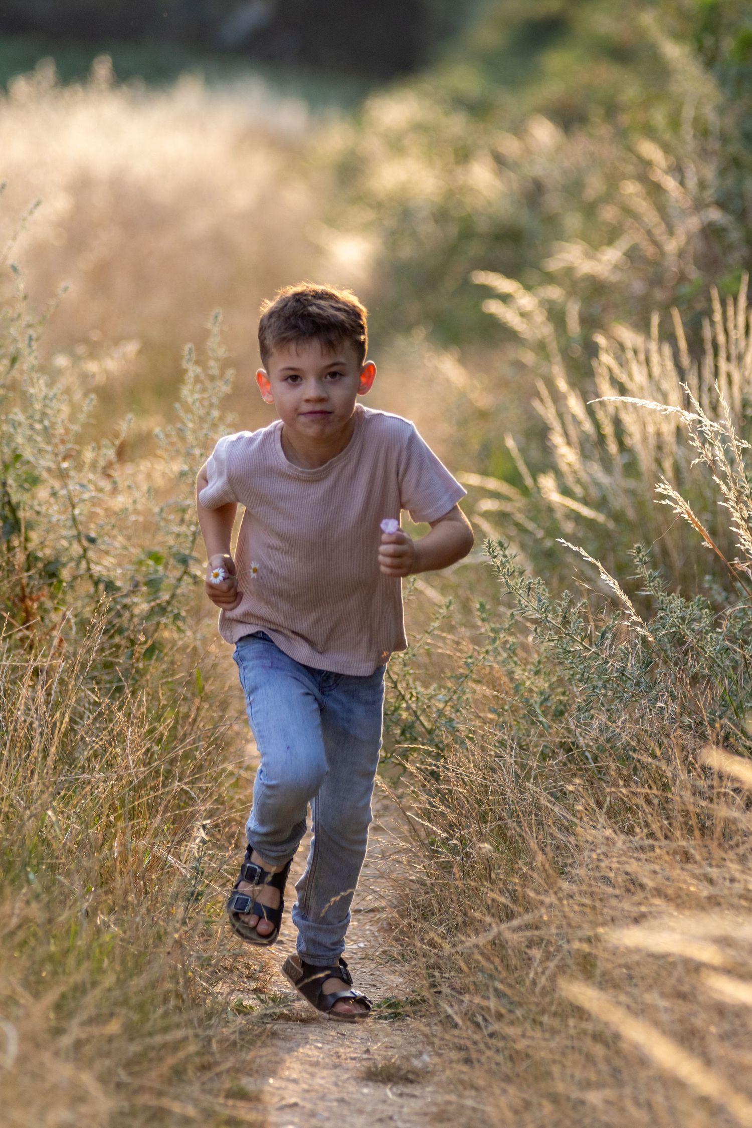 Young boy running along dirt path through tall golden grass in sunlight.