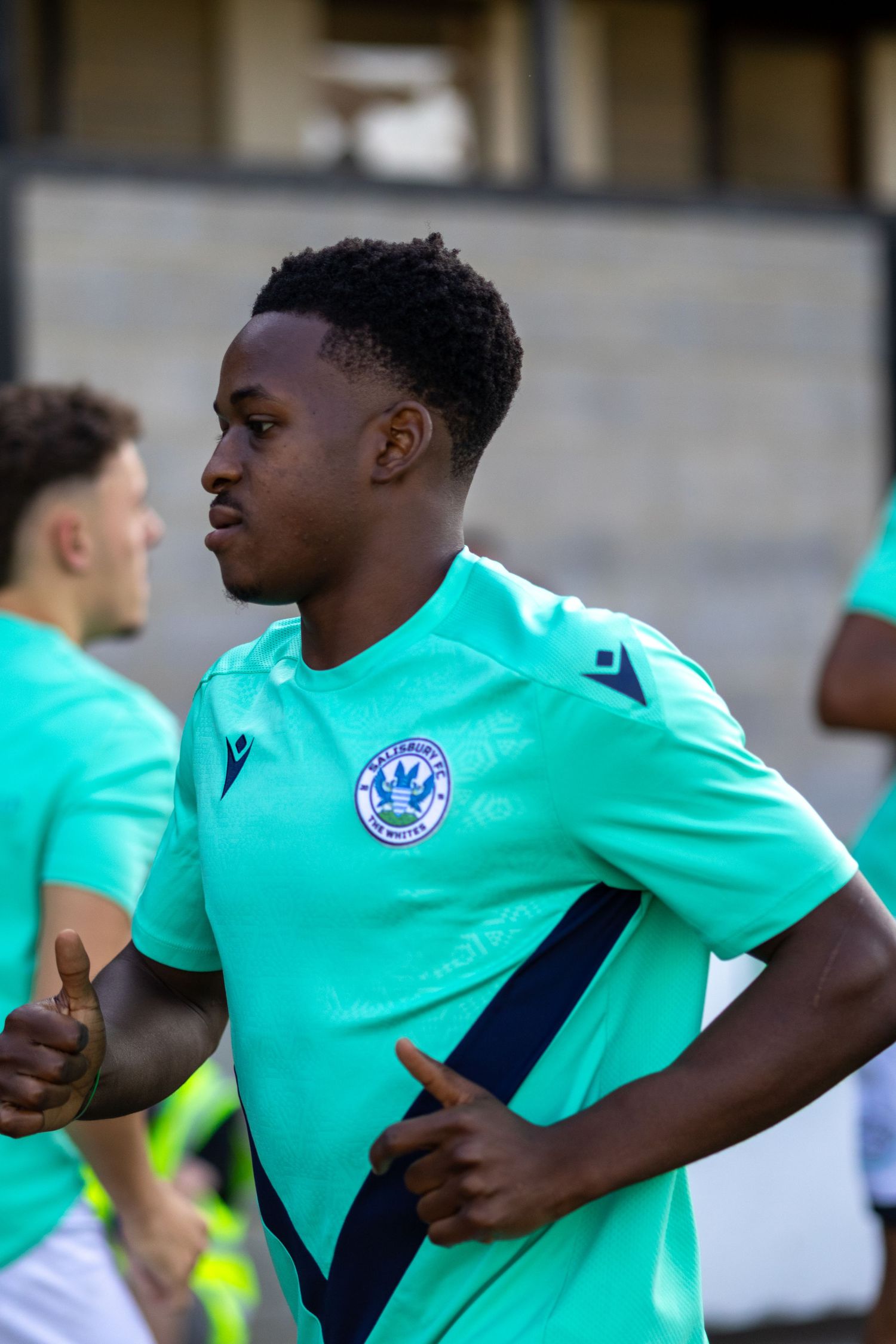 A Black soccer player in a teal green Macron kit jogs during a pre-match warmup session.