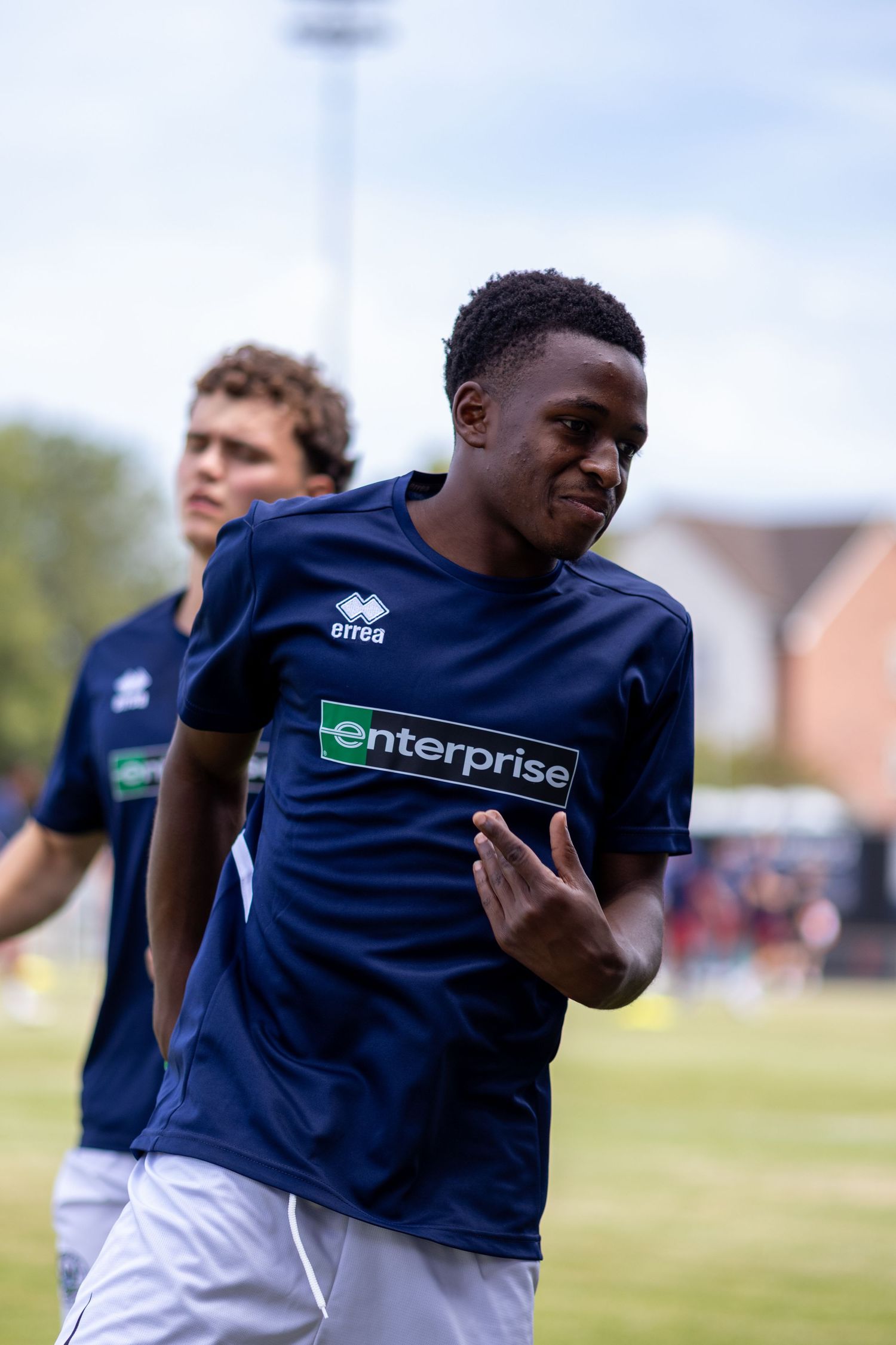 Young Black footballer in navy Enterprise Errea kit jogging during outdoor pre-match warmup session.