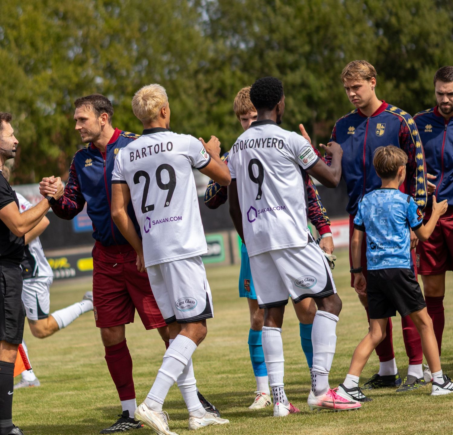 Football players wearing numbers 29 and 9 shake hands with opponents on a sunny grass pitch after a match.