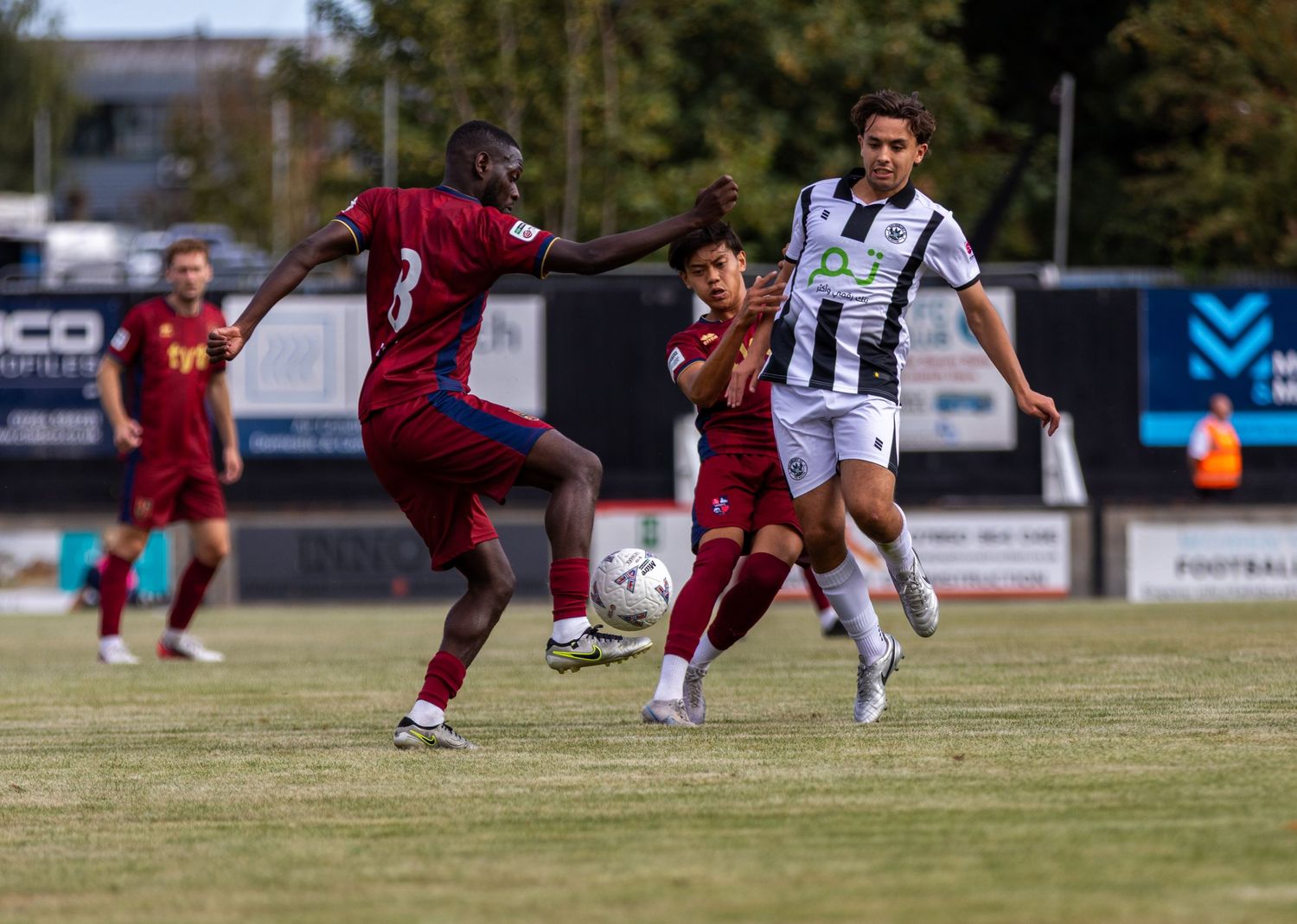 Two soccer players in maroon and one in black and white compete for the ball in a midfield challenge.