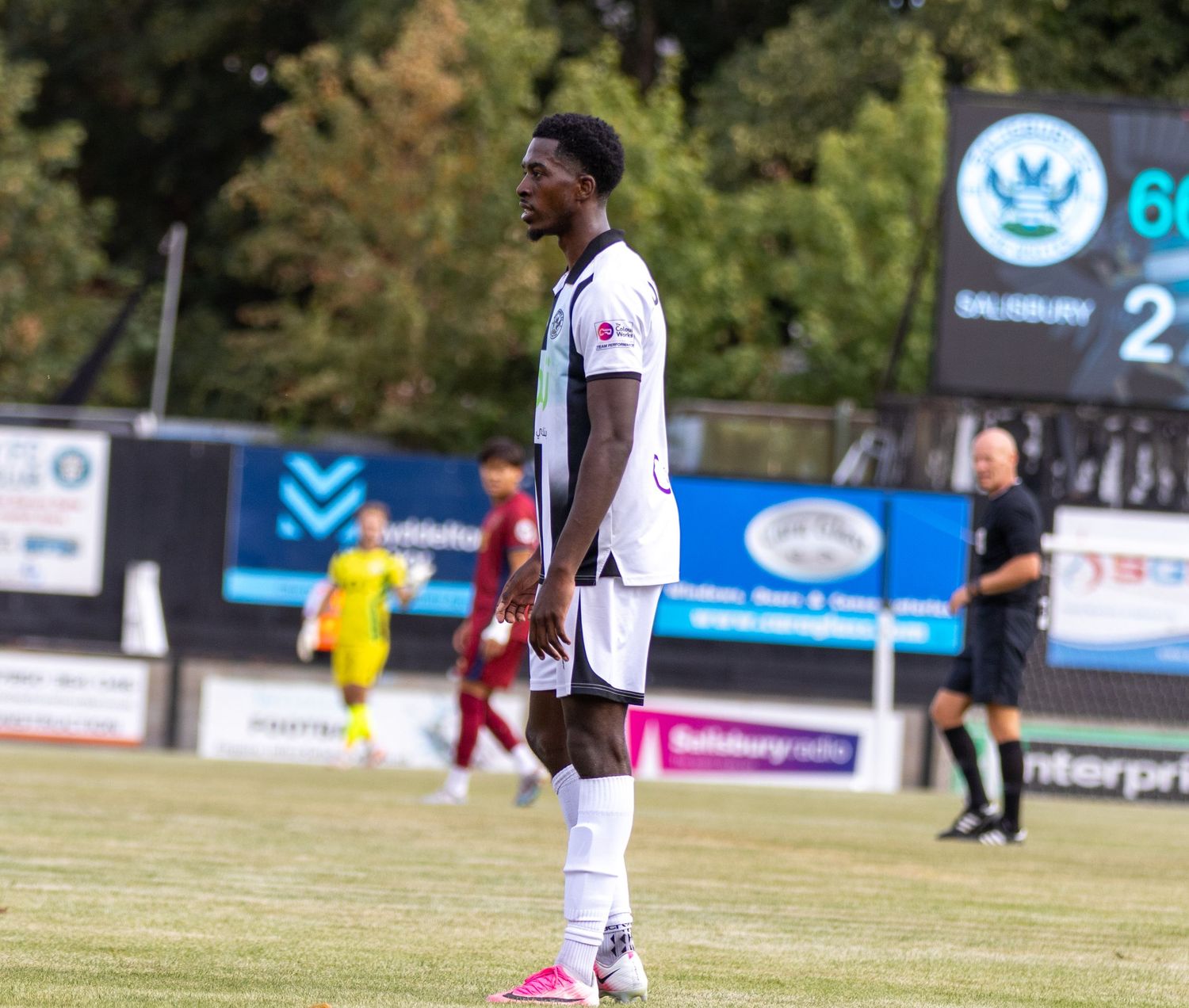 Football player in white kit stands on pitch with scoreboard showing Salisbury 2 in the background.