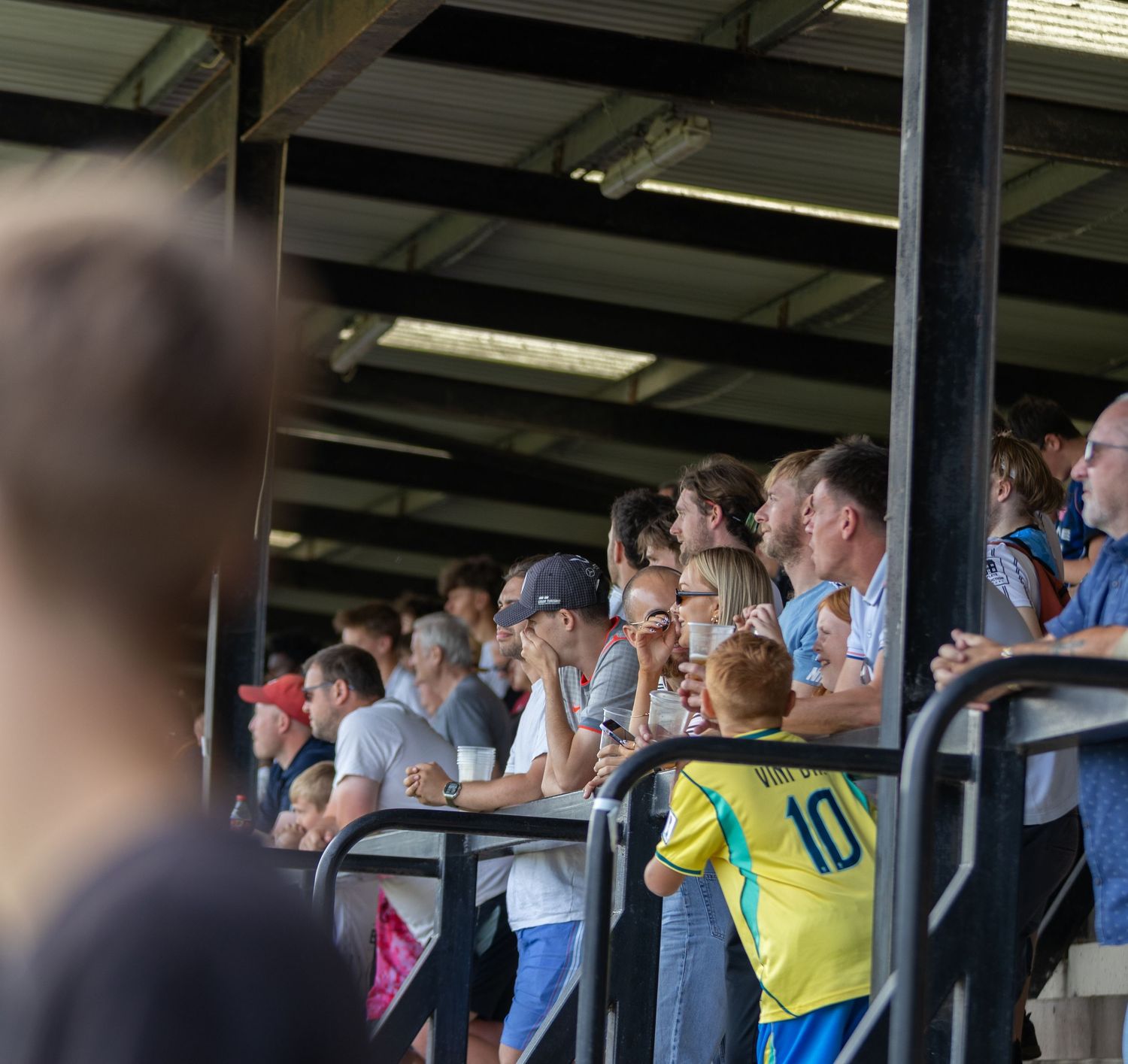 Football fans watch from a covered stand as a young child in yellow kit stands at the barrier.