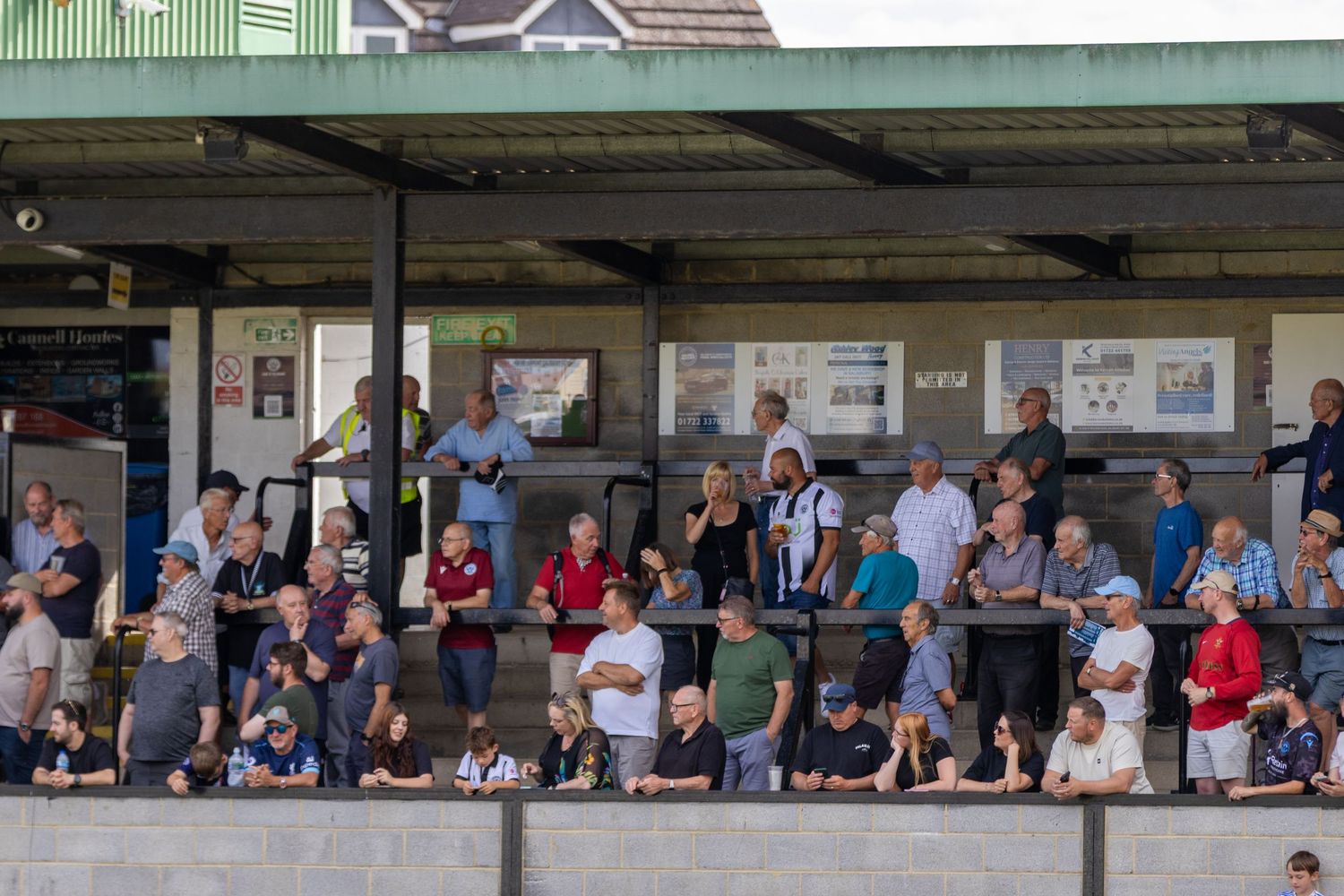 Football supporters stand under a covered terrace watching a match at a non-league ground.