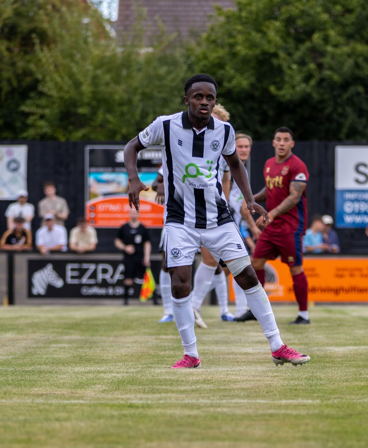 A Black footballer in black and white striped kit with pink boots plays in a match.