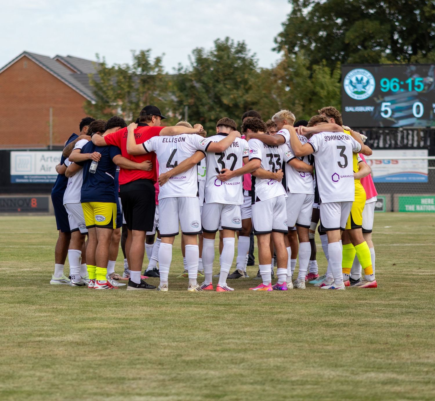Soccer team huddles together at full time with 5-0 scoreboard visible in background.