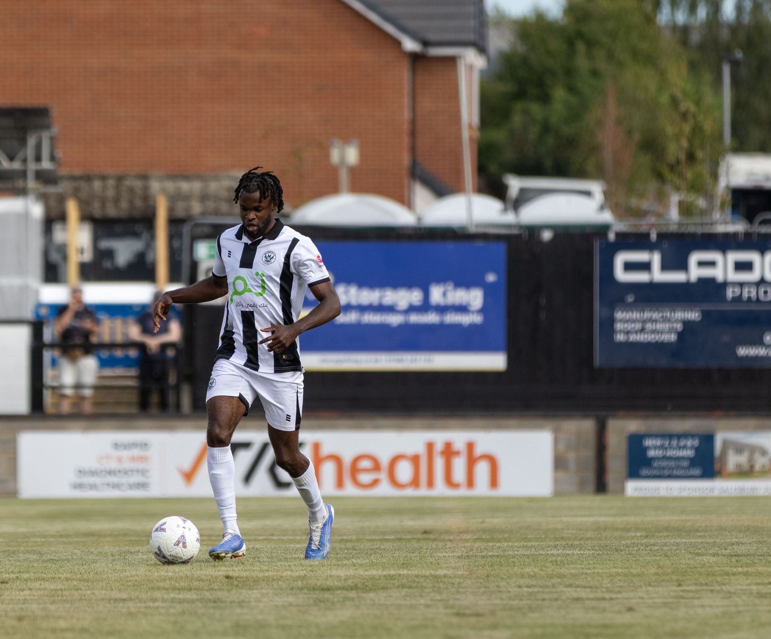 A soccer player in black and white kit dribbles the ball on a sunny outdoor pitch with advertising boards in background.