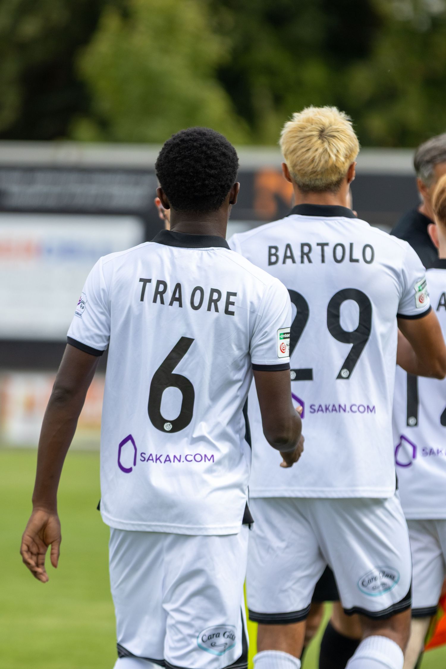 Traore number 6 and Bartolo number 29 in white Sakan.com sponsored football shirts on the pitch.
