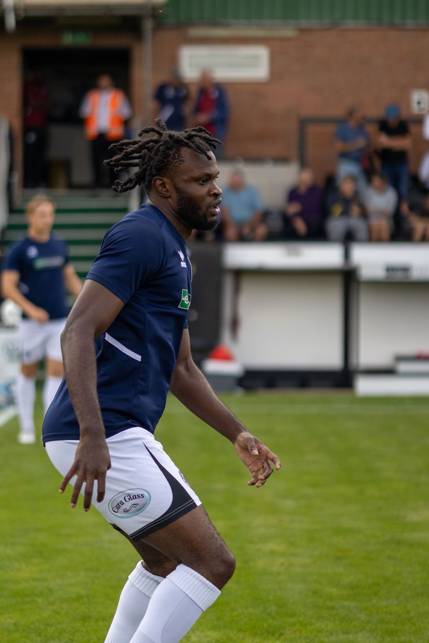 A Black male footballer in navy blue kit moves dynamically on a grass pitch with spectators watching from stands.