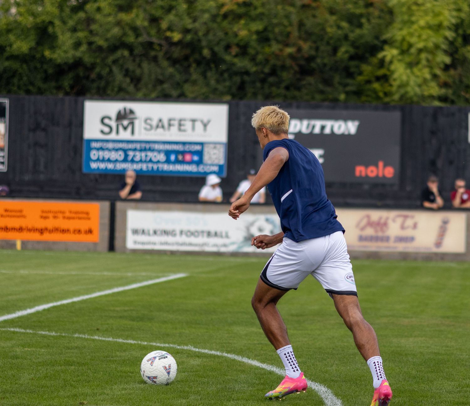 Soccer player with blonde hair in navy blue kit dribbling a ball during warmup at a non-league ground.