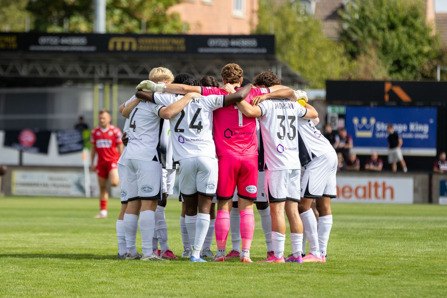Football team in white kits huddle together on sunny pitch before a match with number 33 visible.