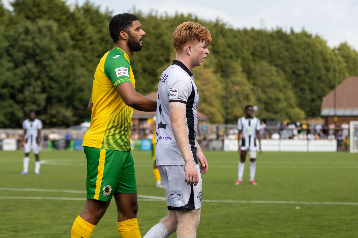Yellow-green kitted player places hand on white-shirted opponent's shoulder during football match.
