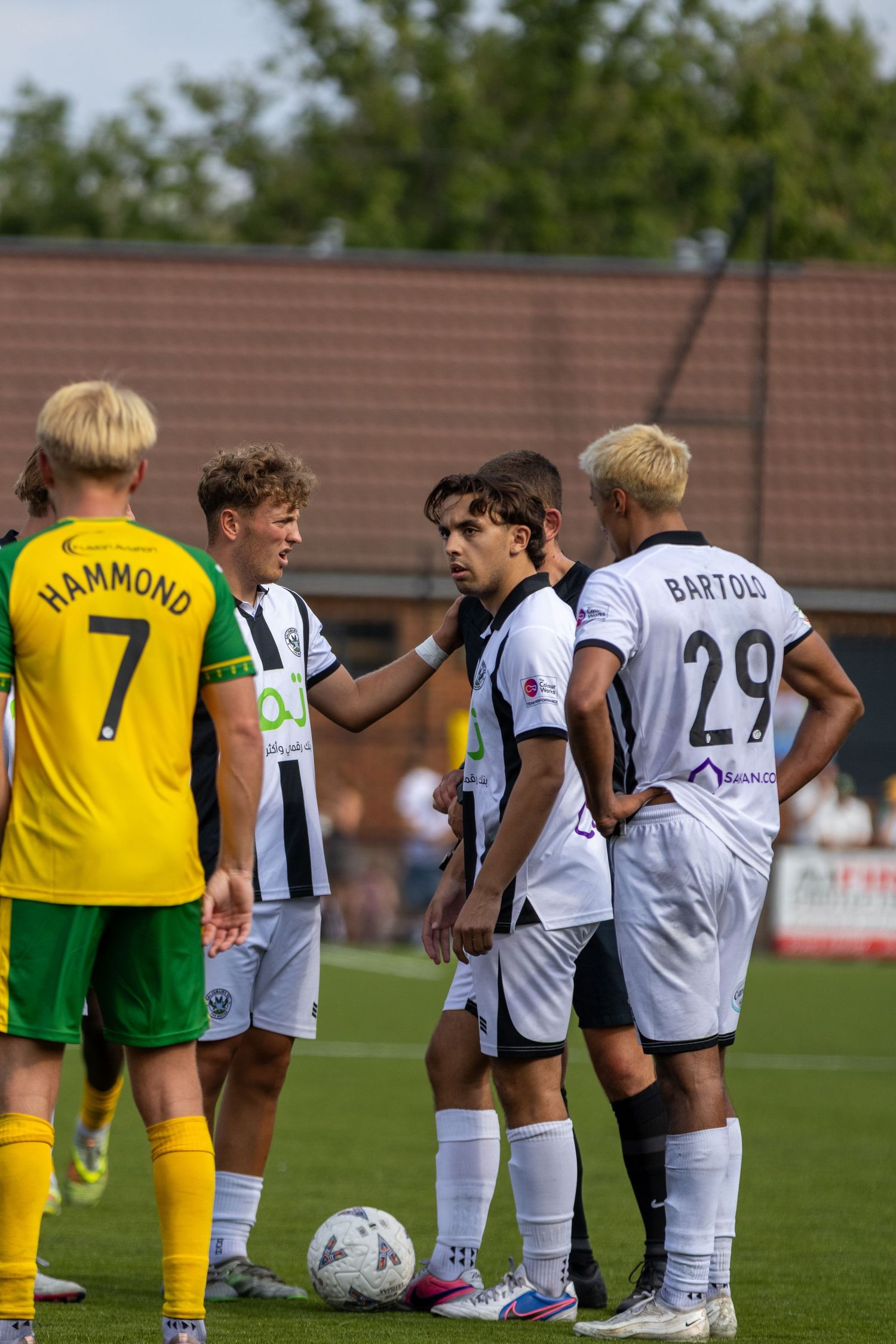 Football players in white and yellow kits huddle near ball including player Bartoli number 29.