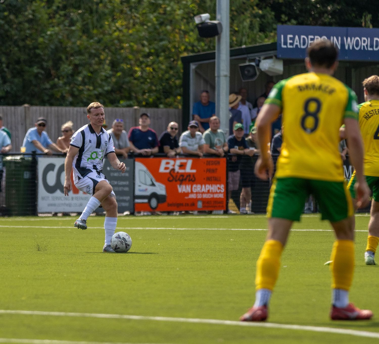 A footballer in black and white stripes passes the ball during a sunny match with a player in yellow kit nearby.