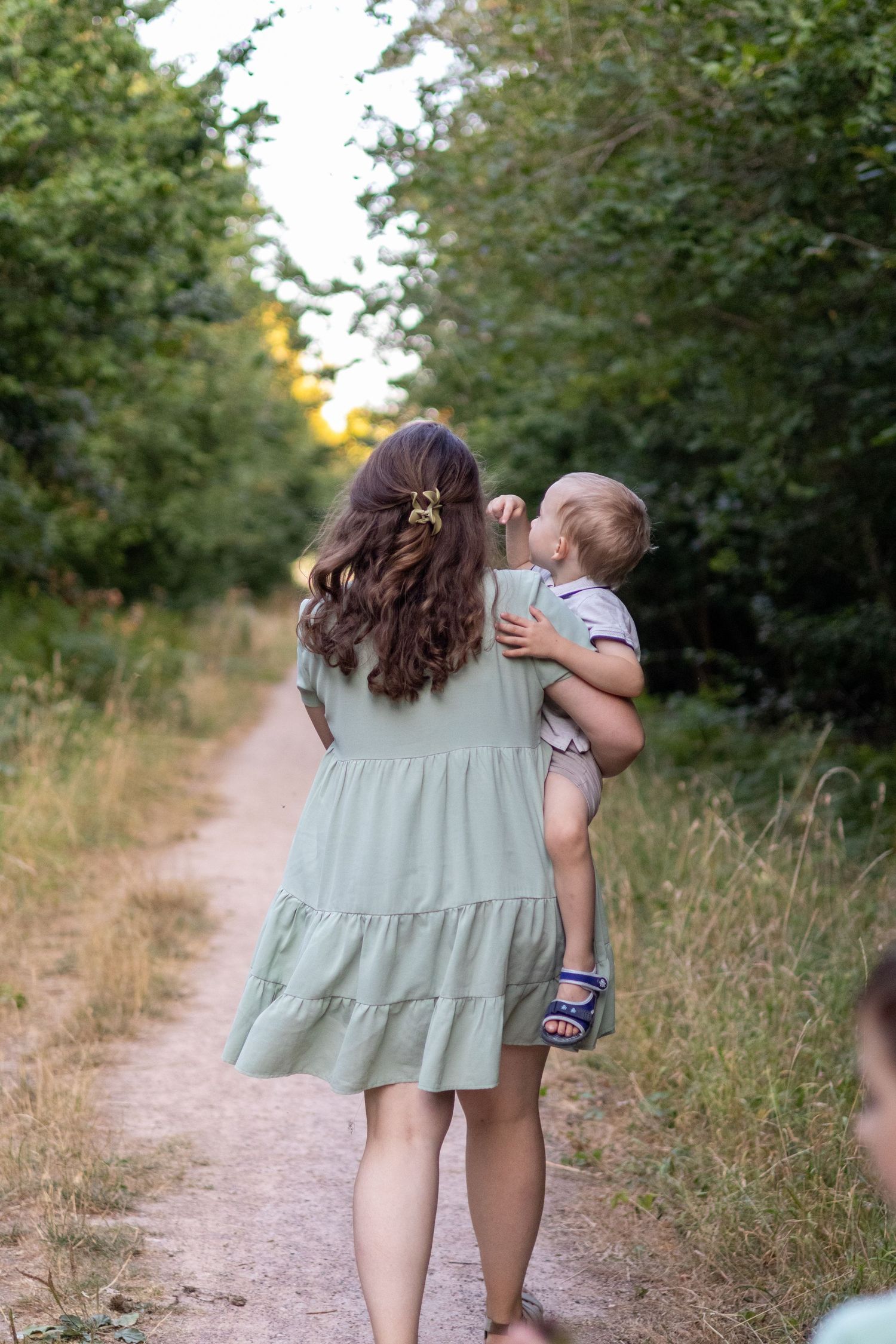A mother in a sage green dress carries her toddler along a tree-lined dirt path.