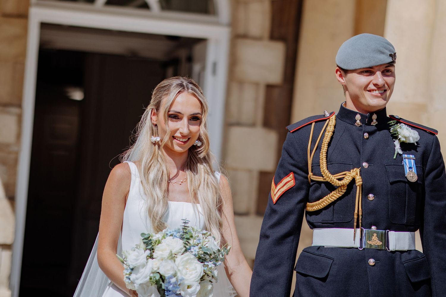 Smiling bride in white gown holding blue and white bouquet walks with groom in military dress uniform.