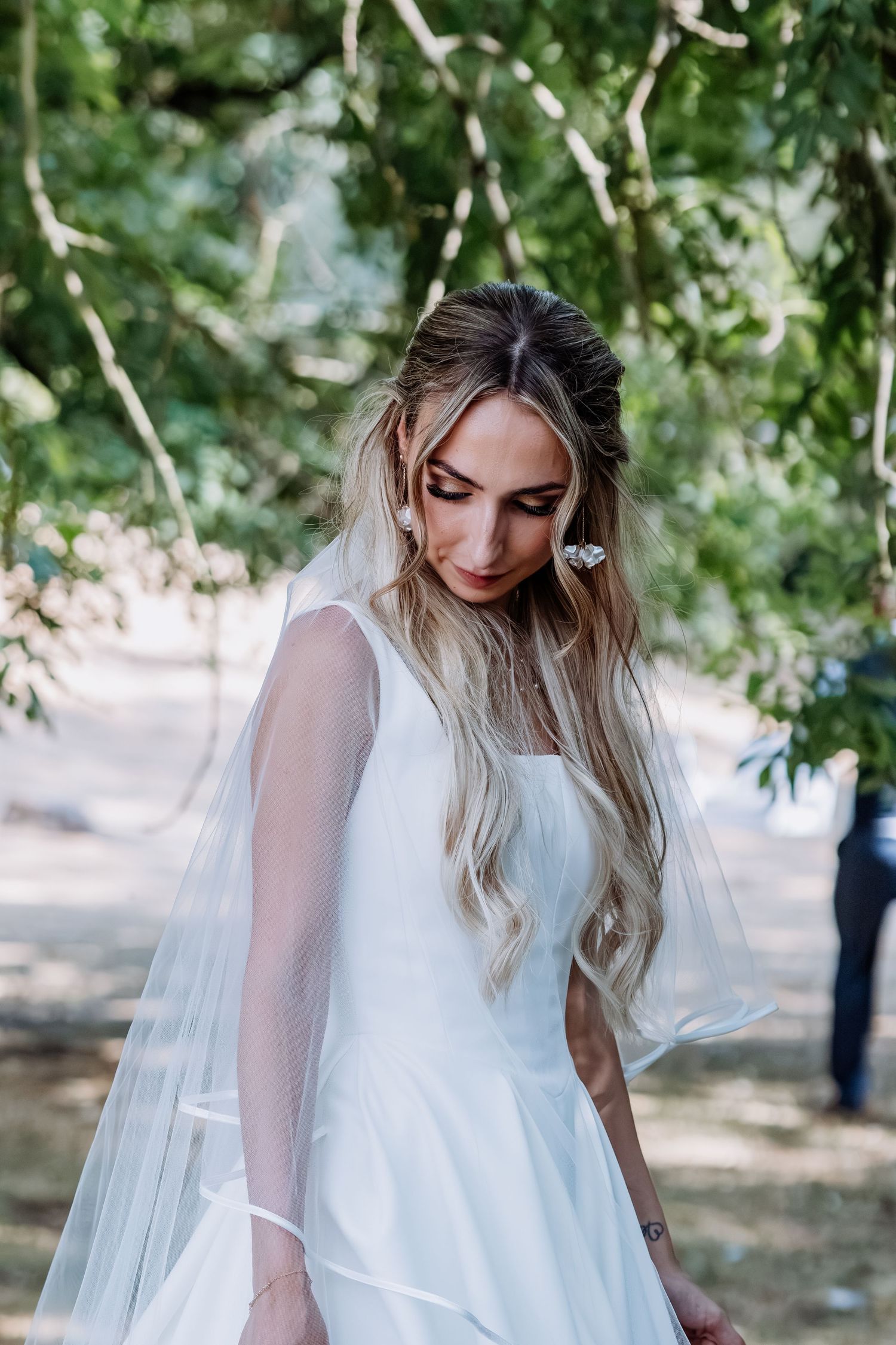 Elegant bride in white gown and veil standing among green trees on her wedding day.