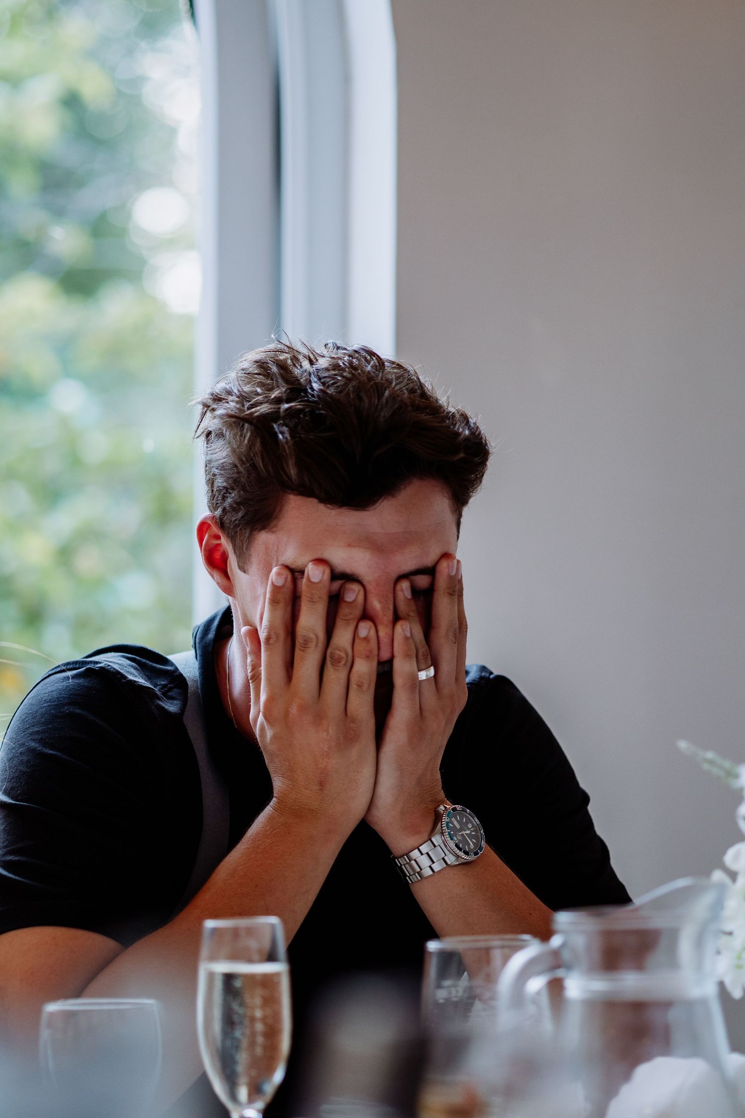 Emotional young man covering face with both hands at wedding reception table with champagne glasses nearby.