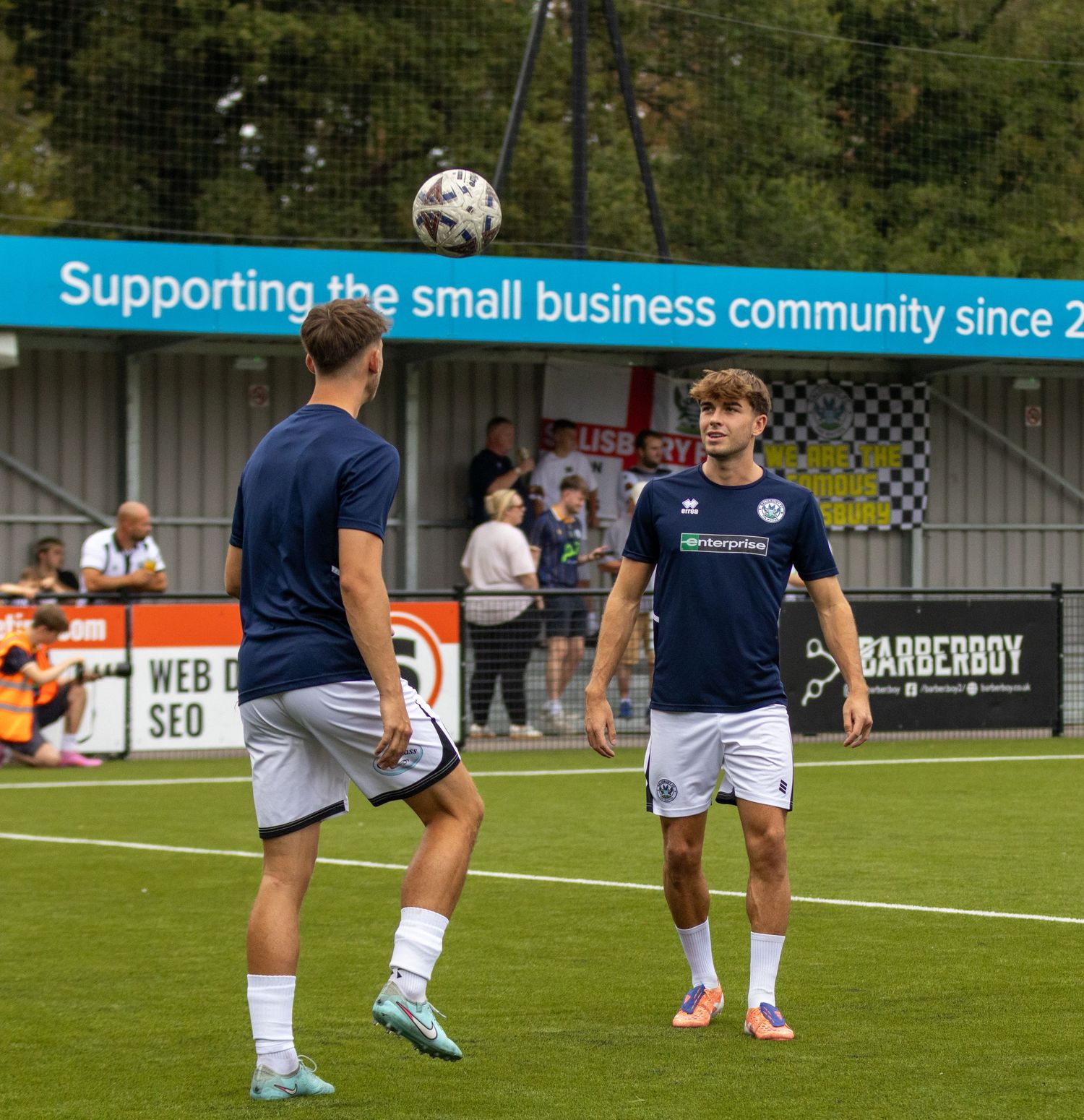 Two footballers in navy kit heading and passing ball during warmup, banner reading supporting small business community.