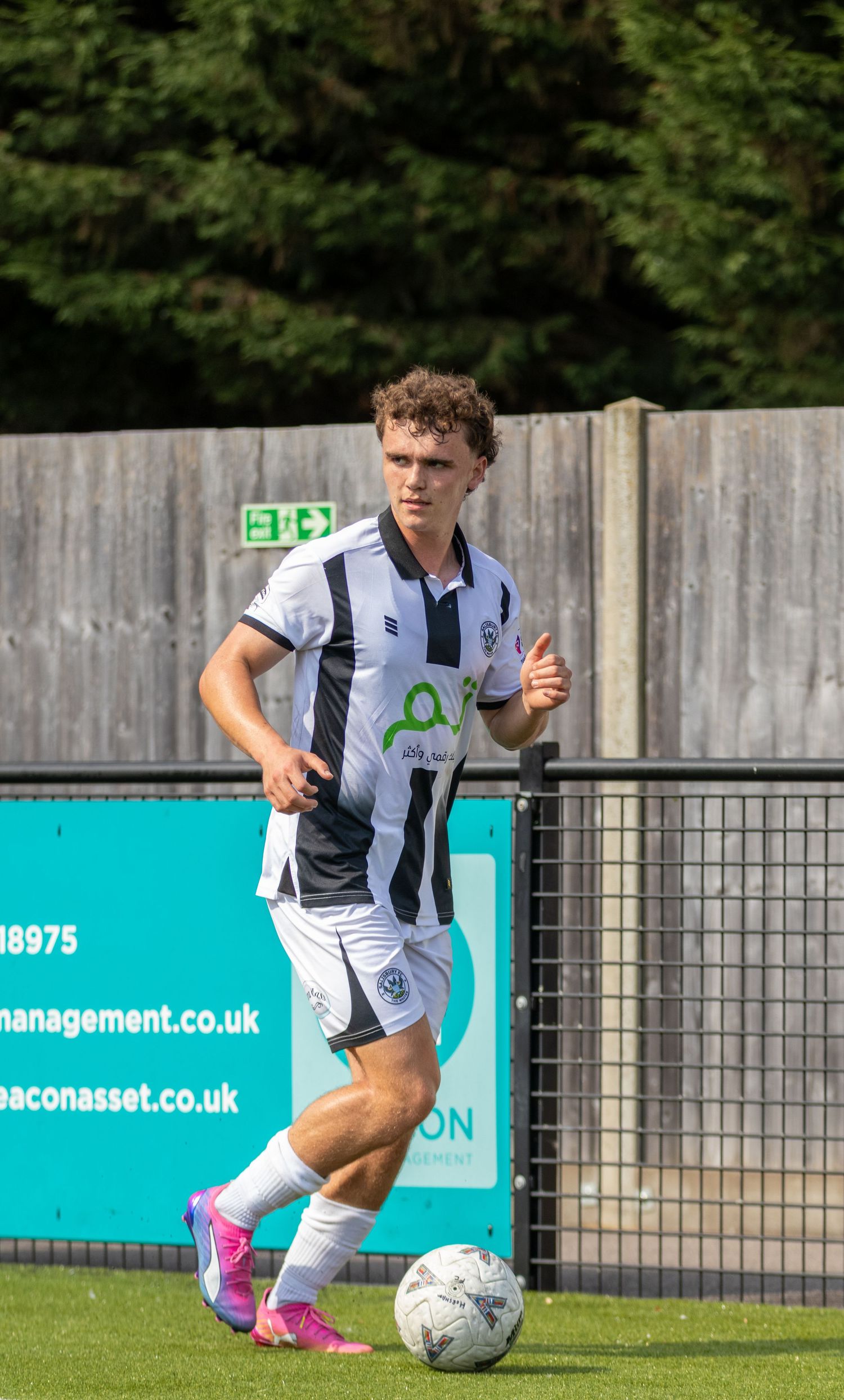 A young footballer in black and white striped kit dribbles a ball along the sideline in bright sunlight.