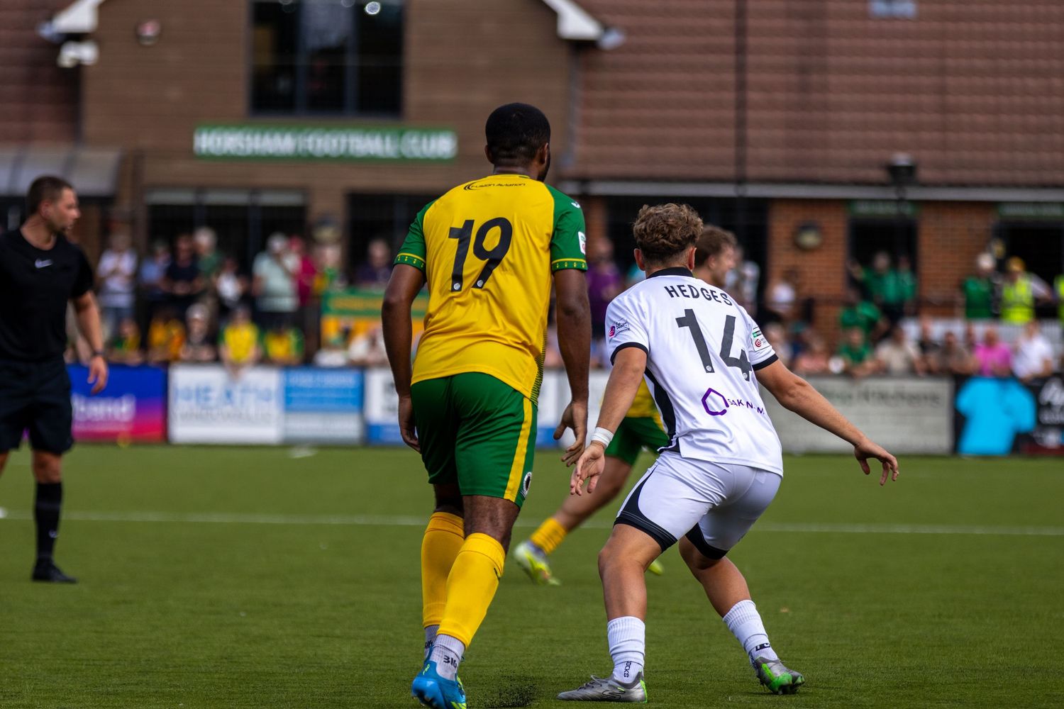 Player number 19 in yellow kit competes against number 14 Hedges in white during a football match.