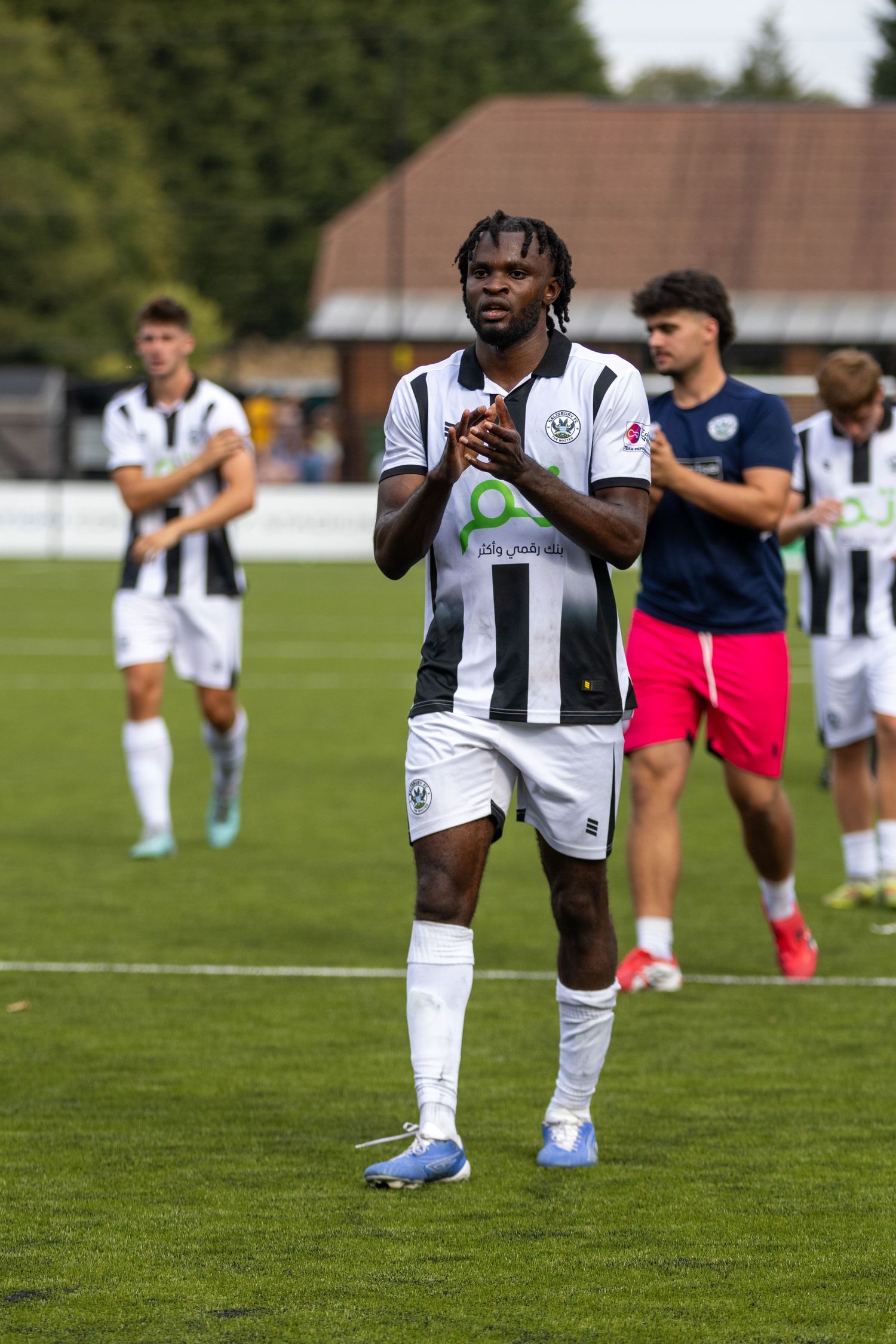 A footballer with dreadlocks wearing black and white striped kit claps on a green football pitch.