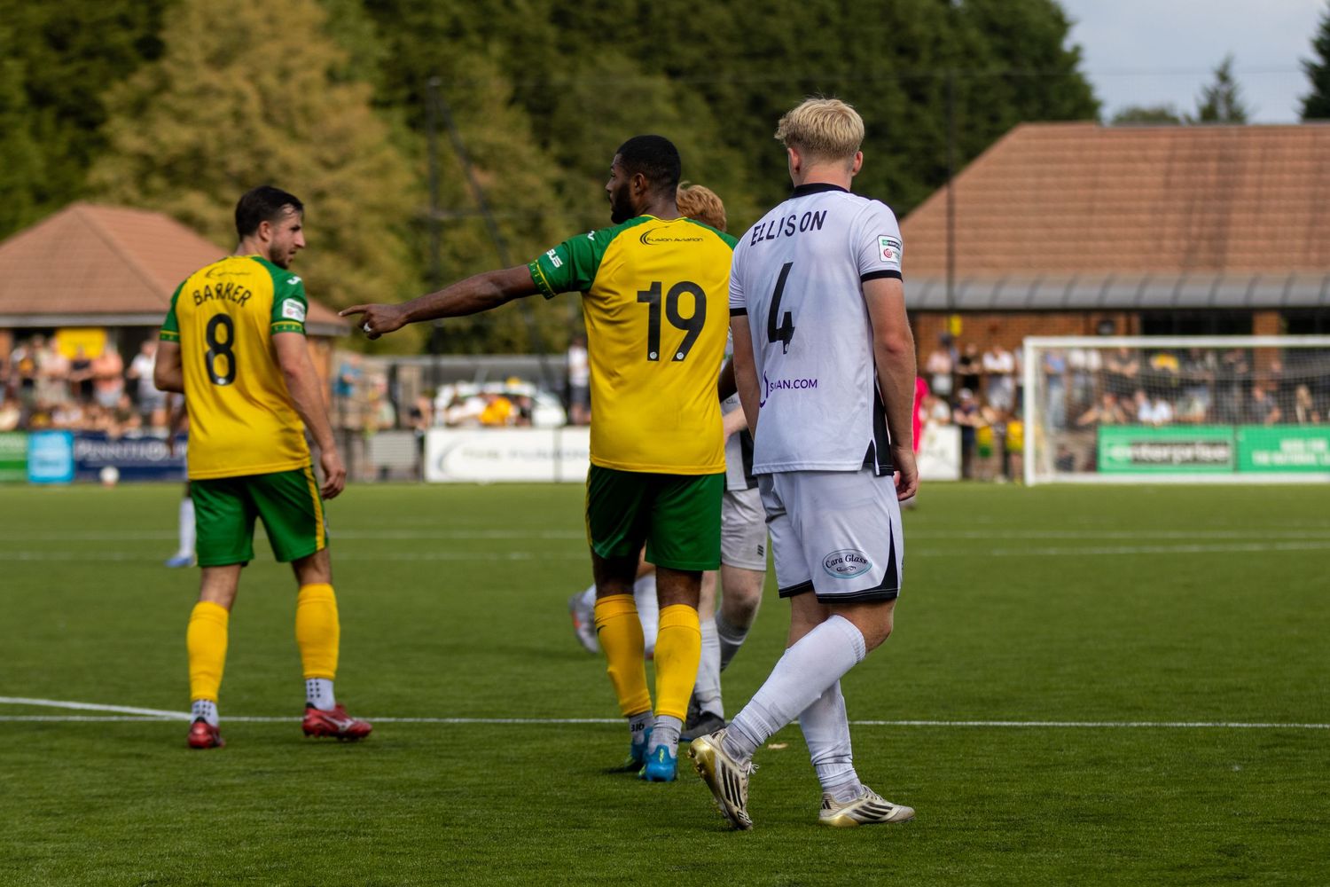 Two yellow-green kit players celebrate near a black-white opponent during a sunny non-league football match.