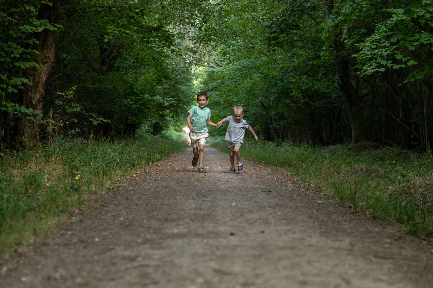 Older child leading younger child by hand running along a scenic green forest trail.
