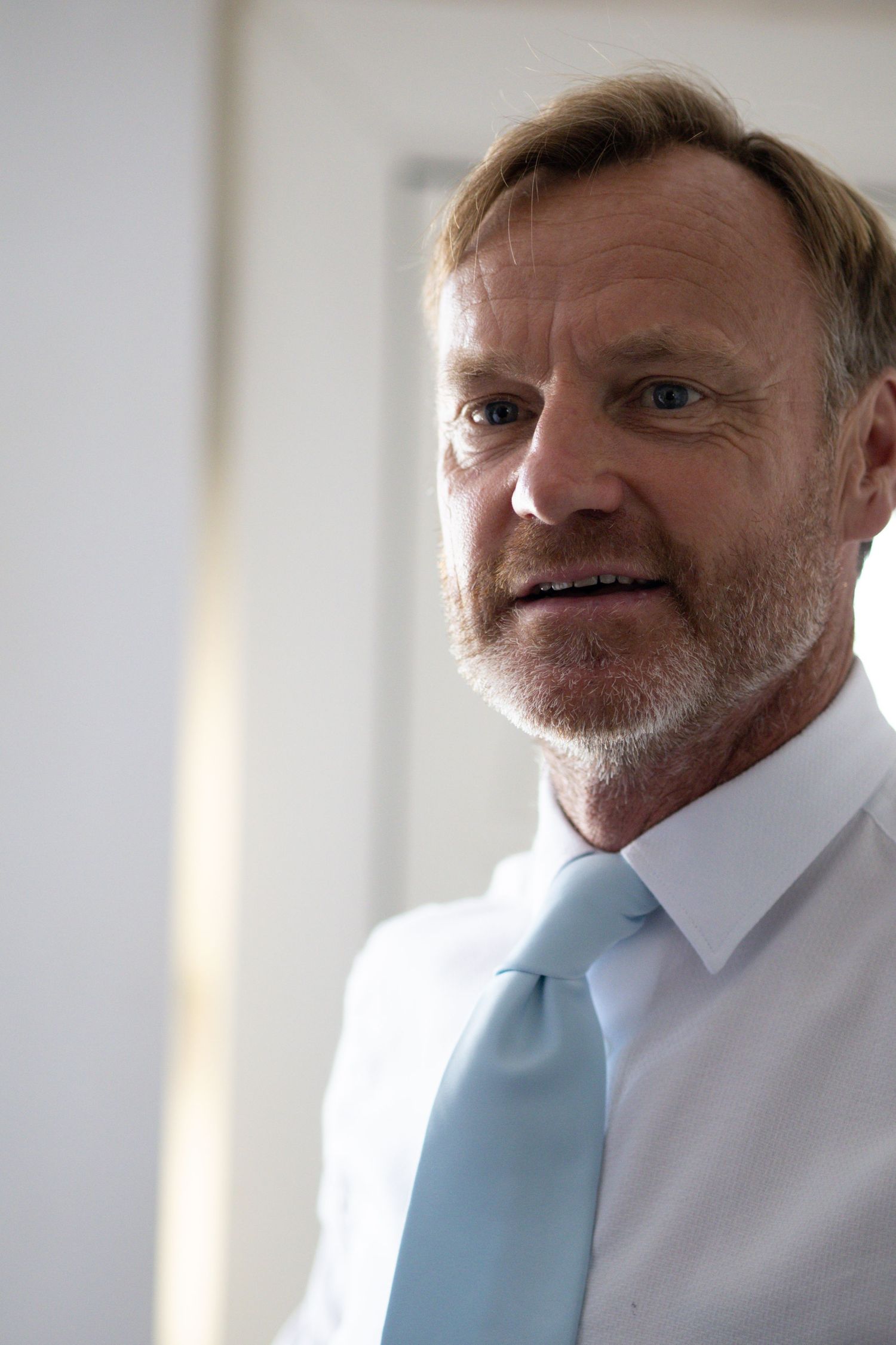 Mature man with salt and pepper stubble wearing white shirt and light blue tie in portrait.