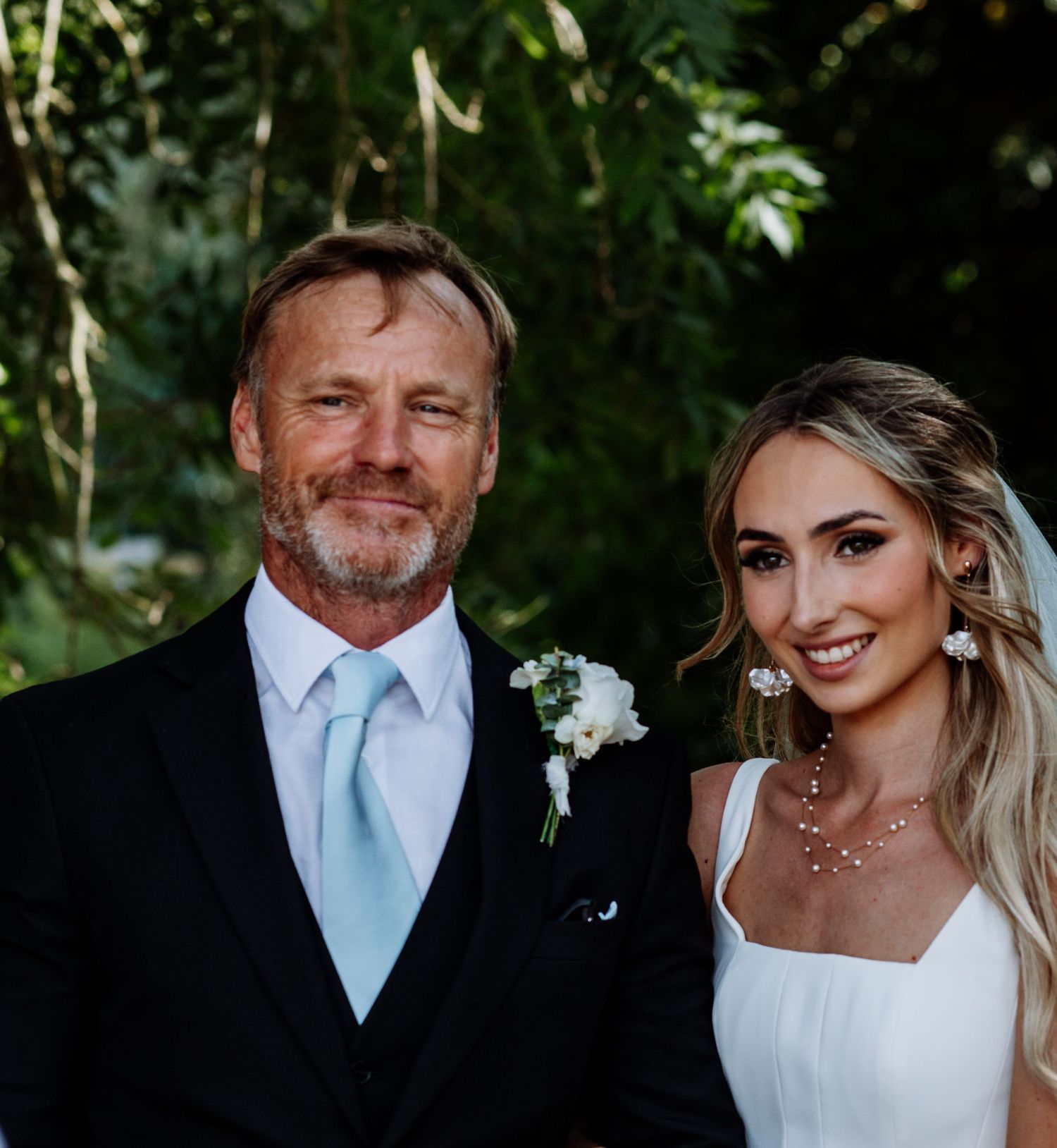 Smiling bride in white wedding dress posing with older gentleman in black suit and blue tie.