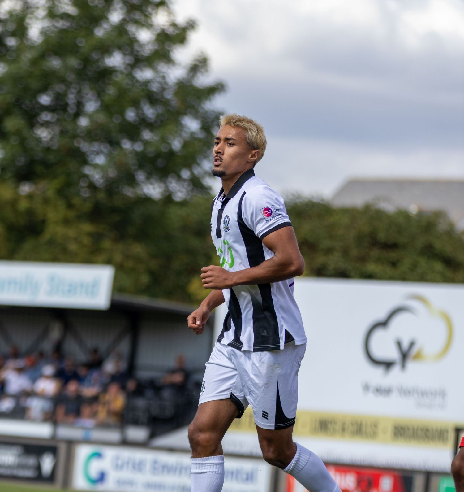 Blonde footballer in black and white strip jogs on the pitch during a sunny football match.