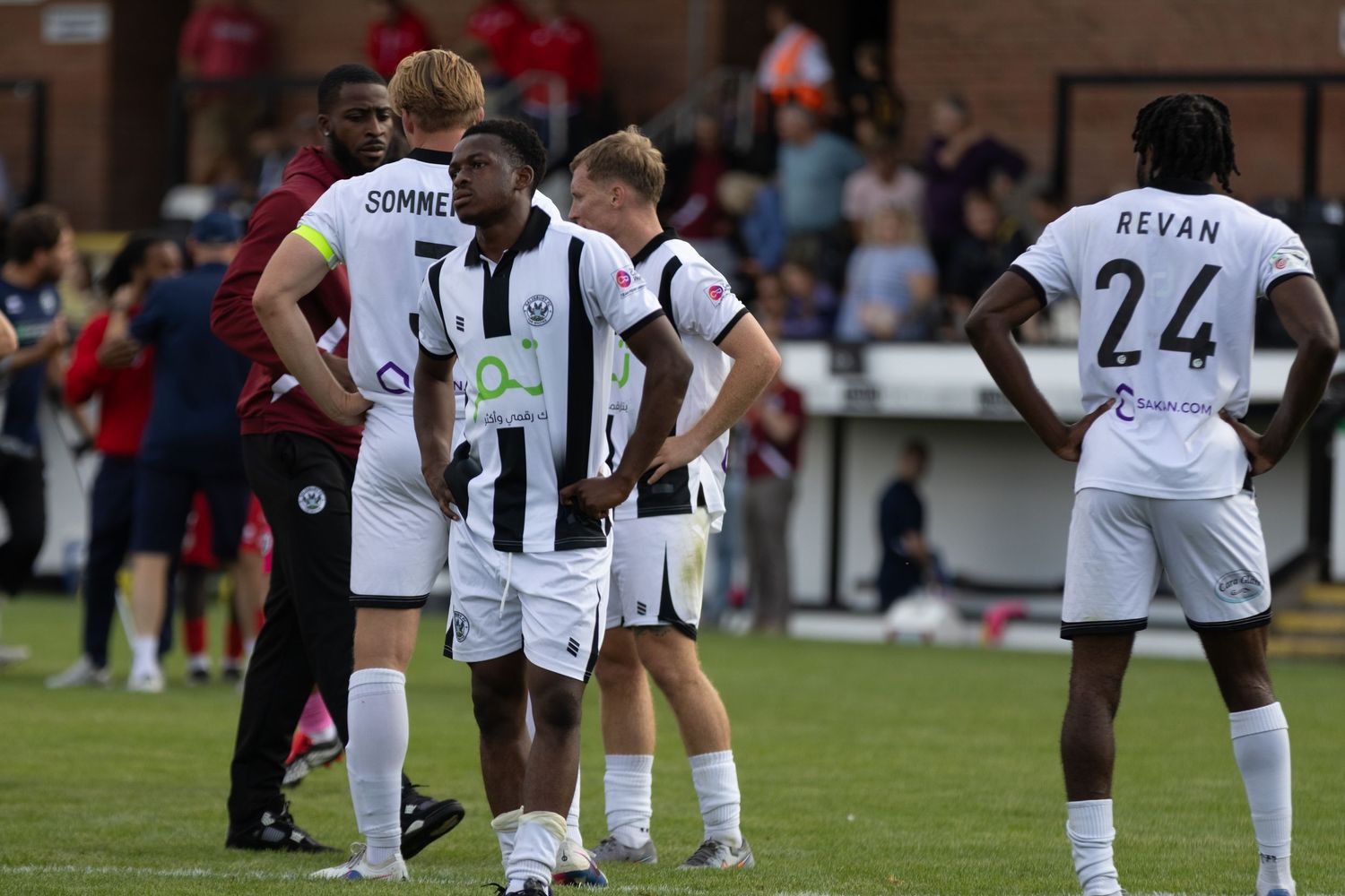 Soccer players in black and white striped jerseys, including Sommerton and Revan, huddle on a grass field during a match.