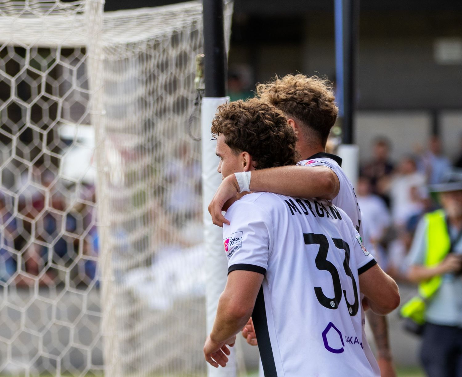 Two soccer players in white uniforms embrace near the goal net during a match.