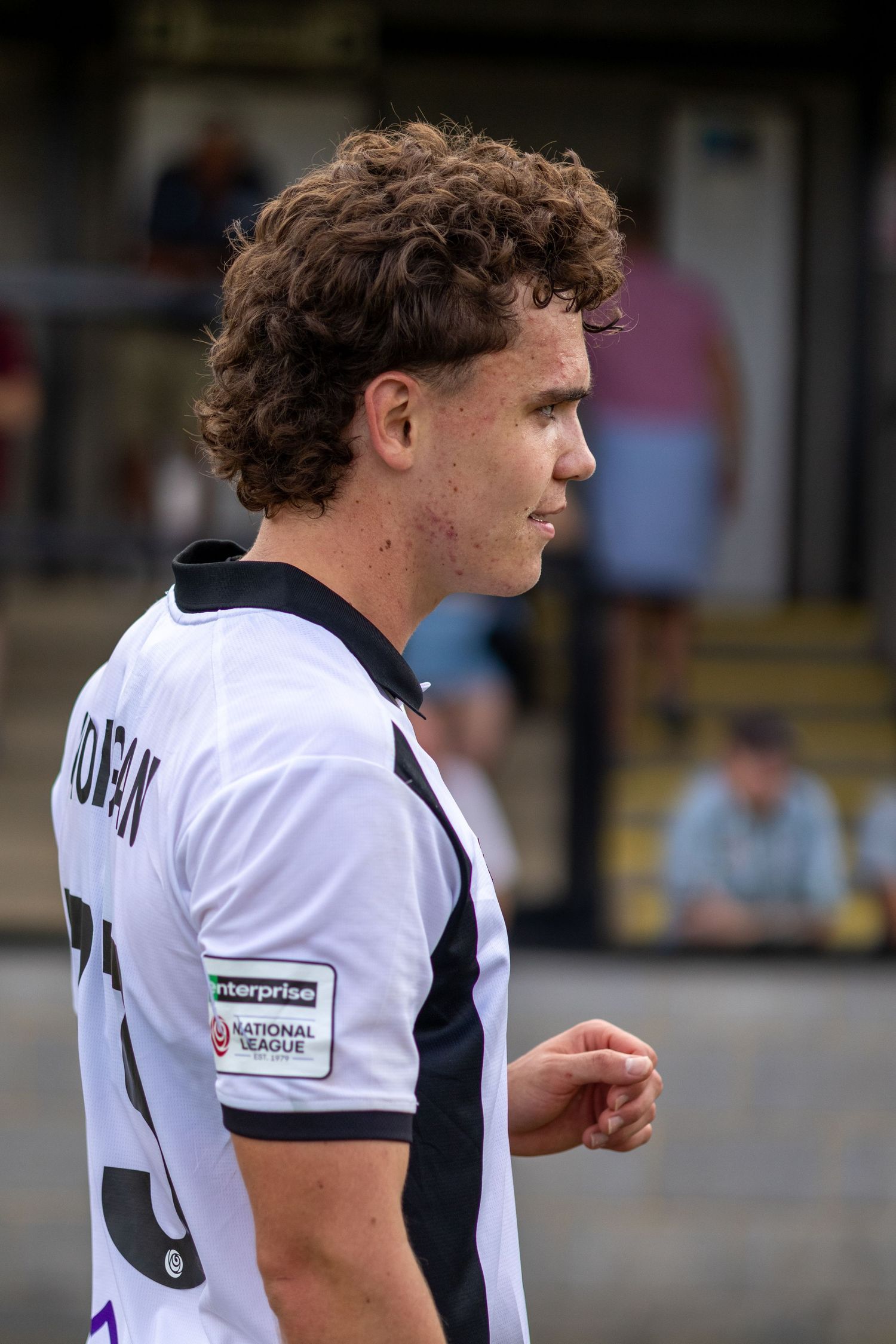 Curly-haired footballer in white National League jersey with enterprise sponsor walks on pitch.