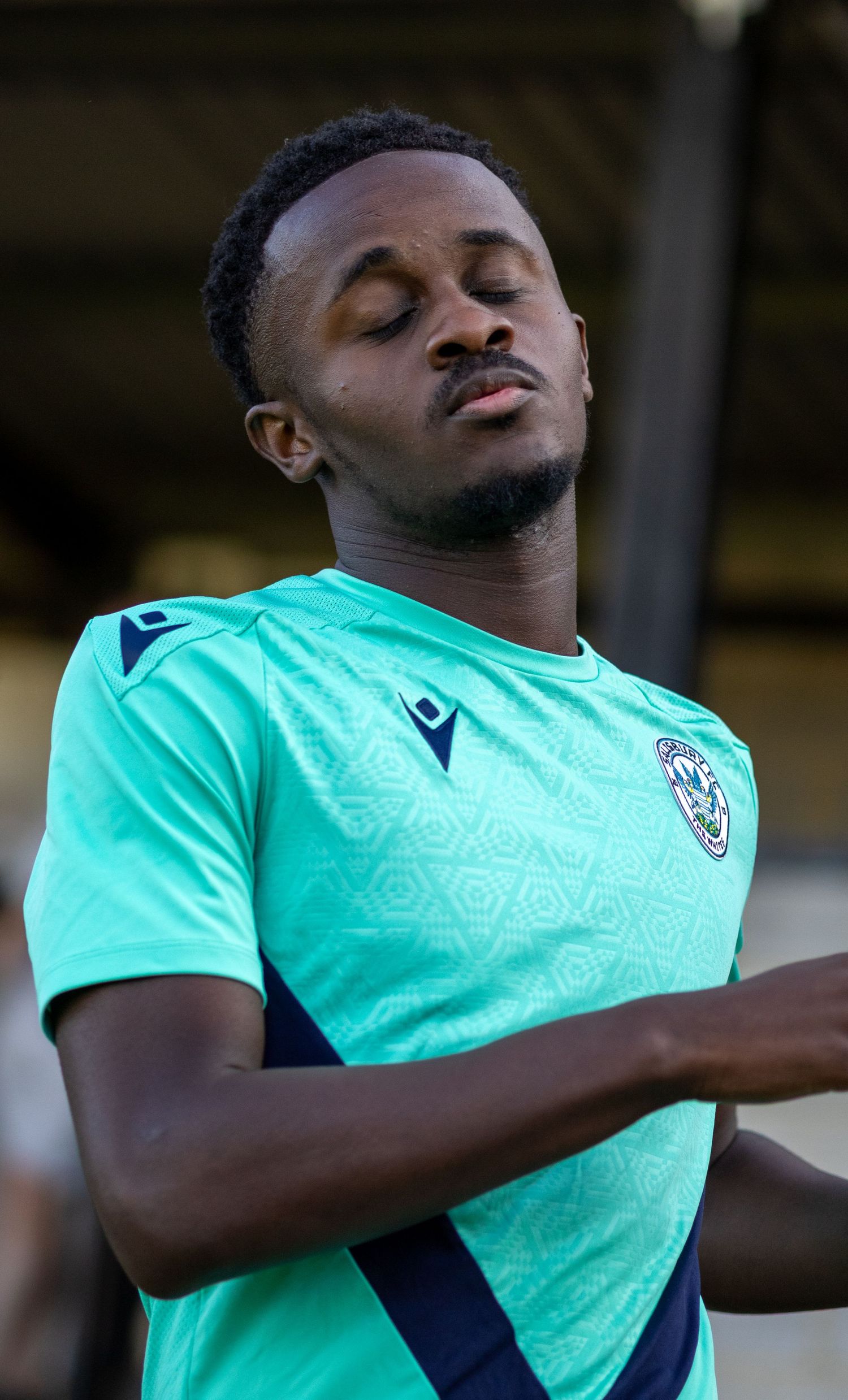 A Black soccer player in a teal green Macron jersey stretches with eyes closed before a match.