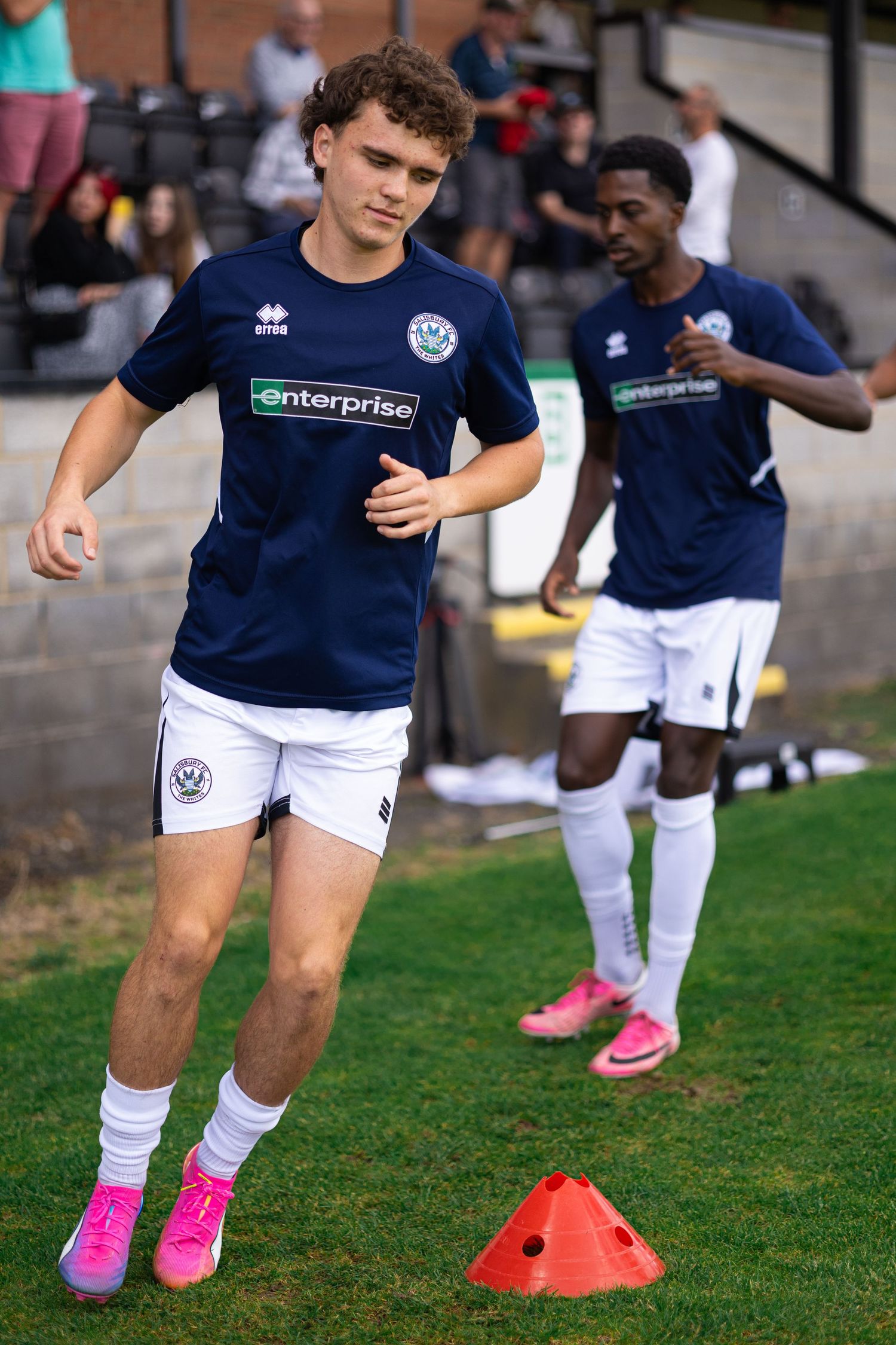 Two soccer players in navy Enterprise-sponsored kits warm up with cones on a sunny outdoor pitch.