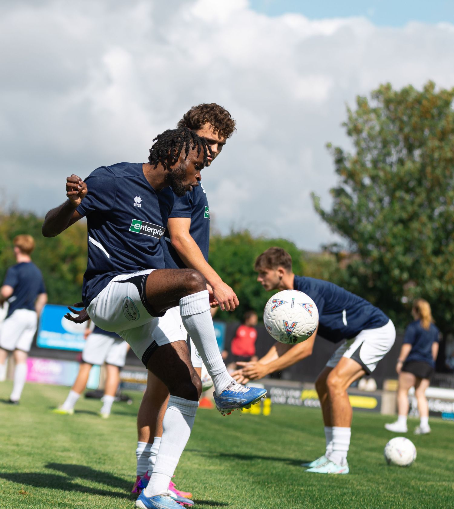 Football players in navy kits warm up on a sunny pitch, with one player controlling a ball during pre-match training.