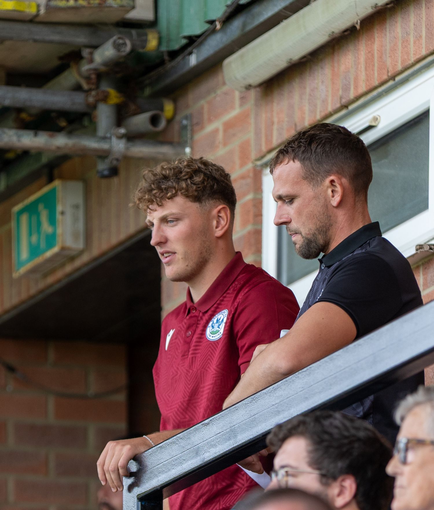 Two men in casual attire stand on a stadium balcony watching a football match on a sunny day.