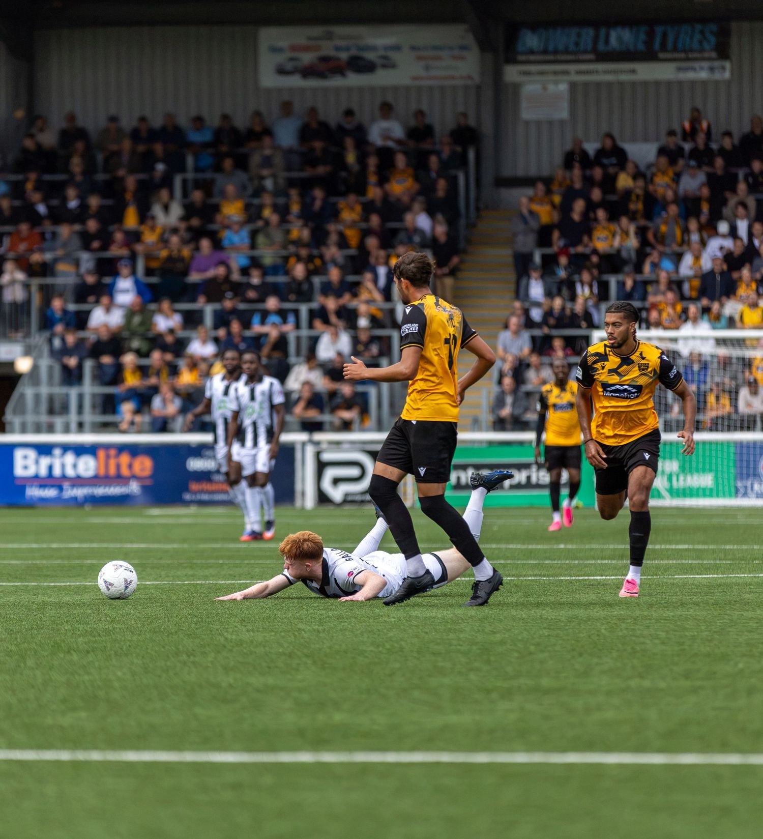 Football match action with player fallen on pitch, others in gold and black kits, fans in stadium stands behind.
