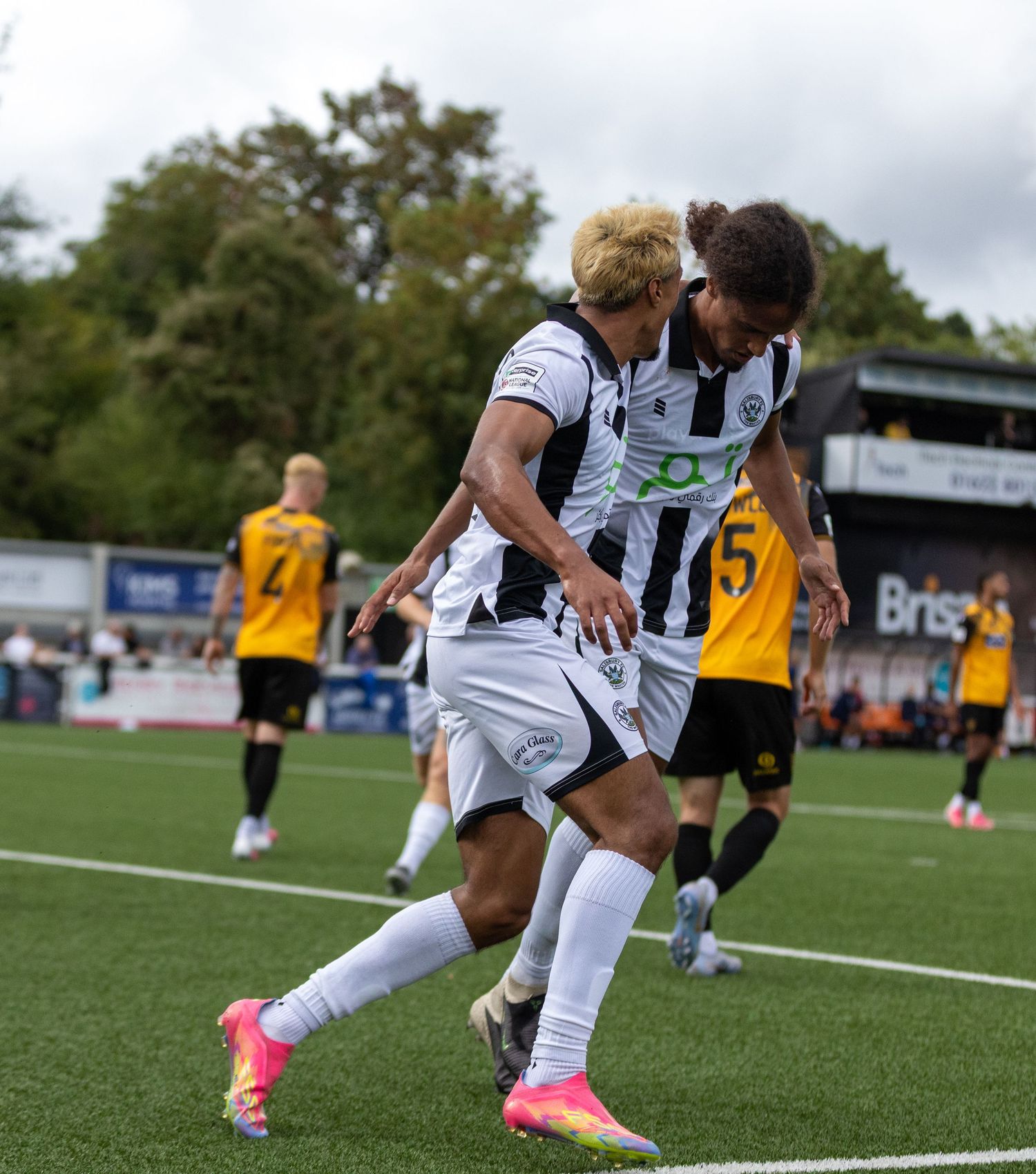 Two football players in black and white kits celebrate a goal during a match, with opponents in yellow visible behind them.